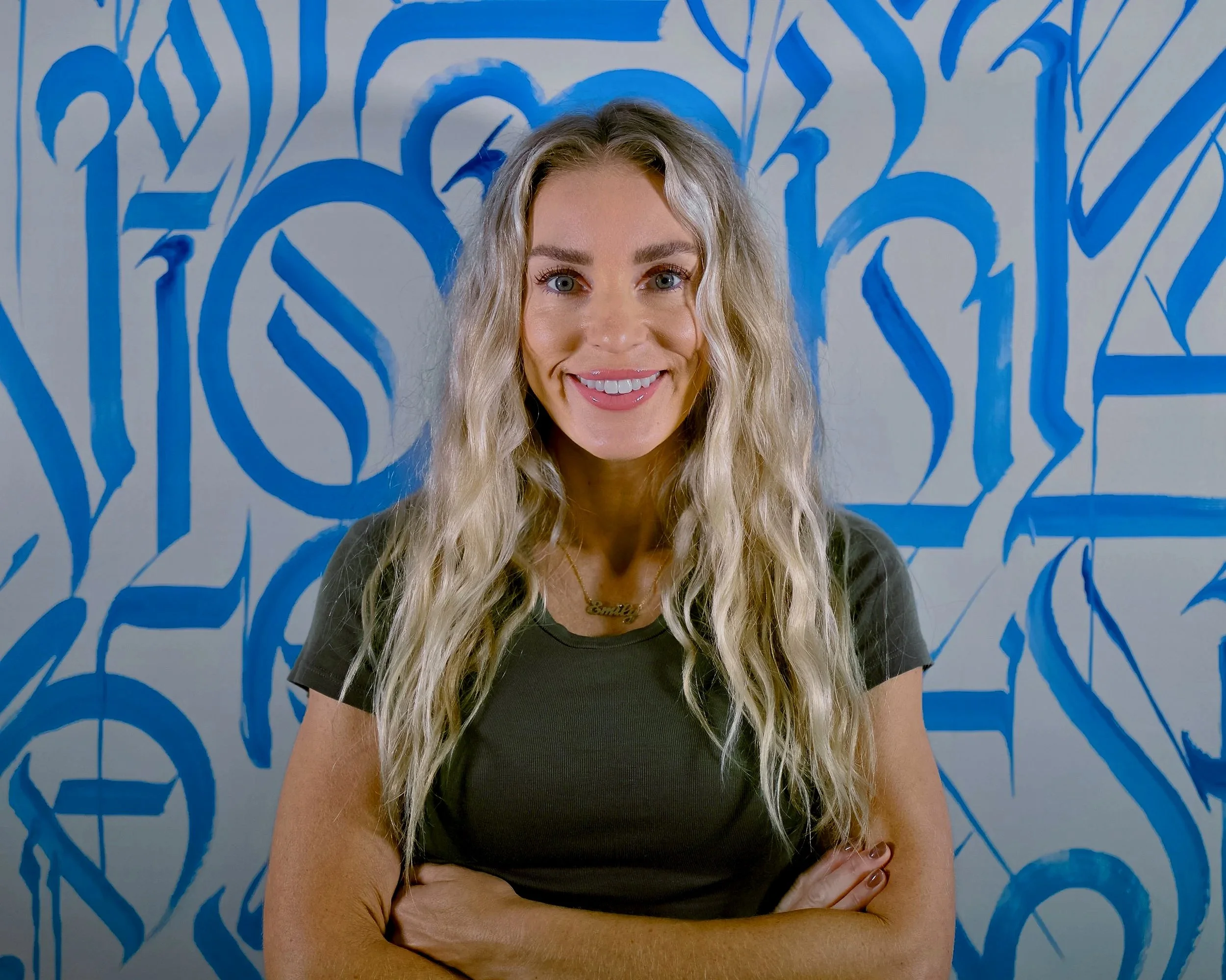A woman with long blonde wavy hair smiling with arms crossed in front of a blue graffiti wall.