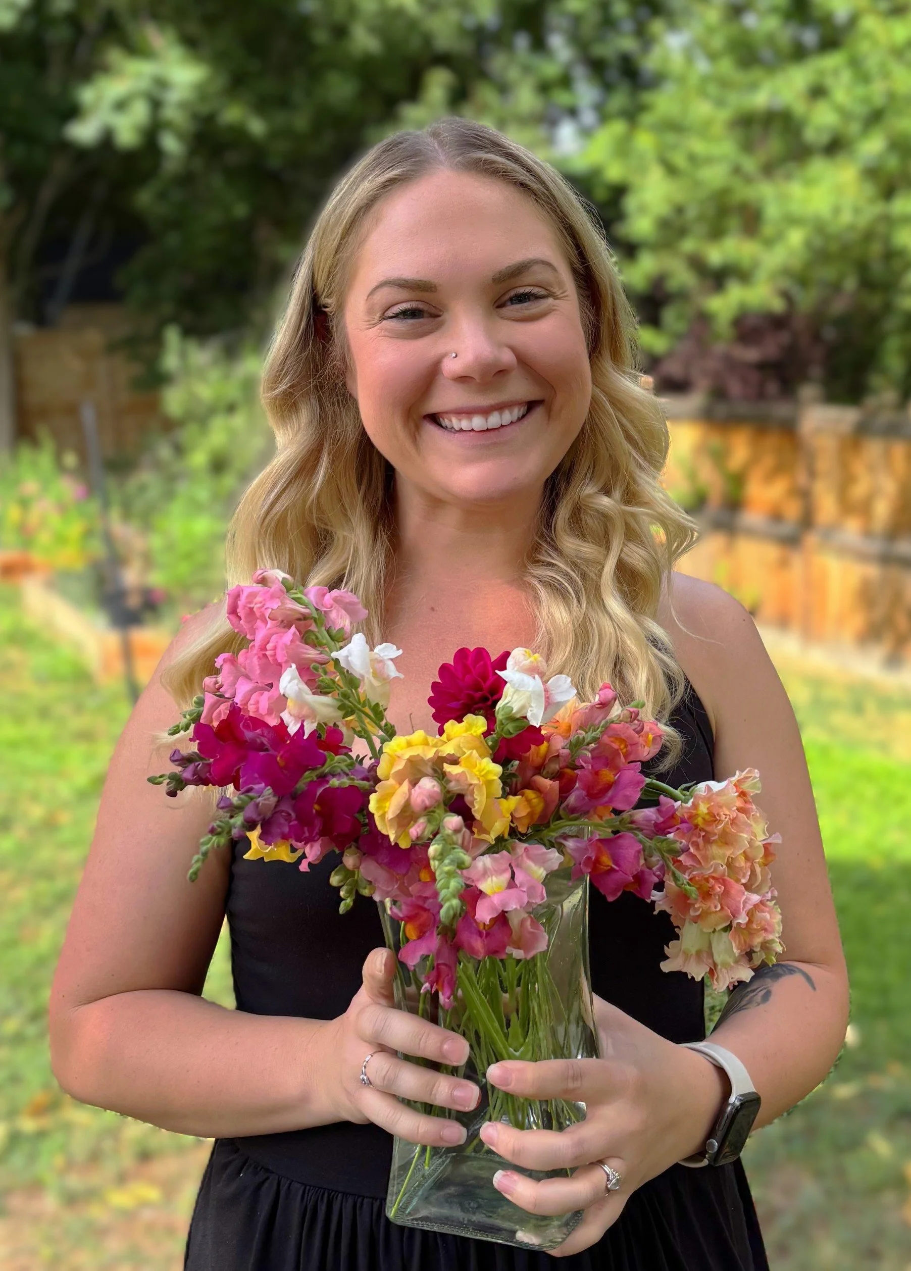 A young woman with long wavy blonde hair smiling and holding a clear glass vase filled with colorful mixed flowers, standing outdoors in a garden with greenery and a stone wall in the background.