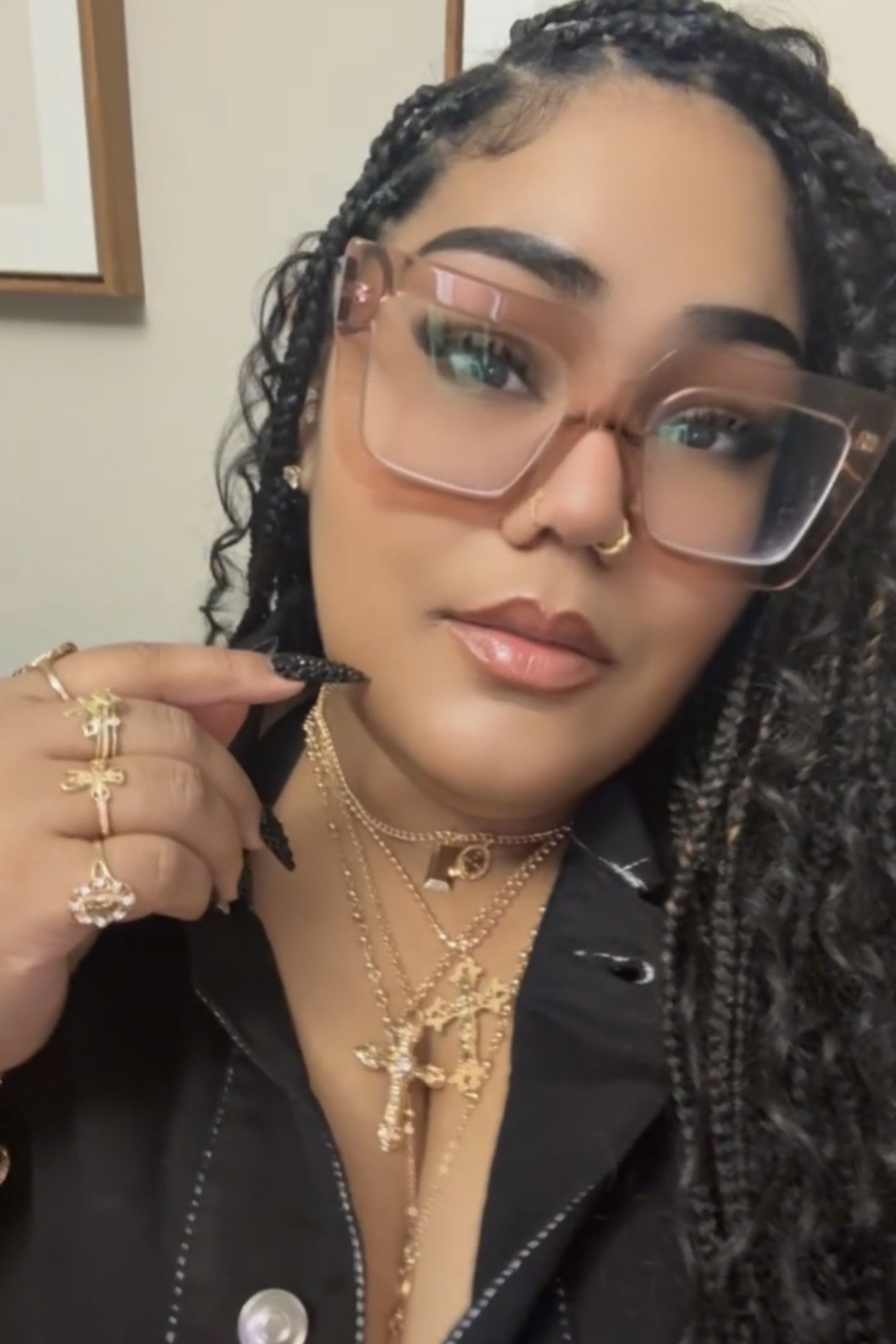 A woman with dark curly hair, wearing large pink glasses, gold jewelry, and a black top, holding her chin with her hand.