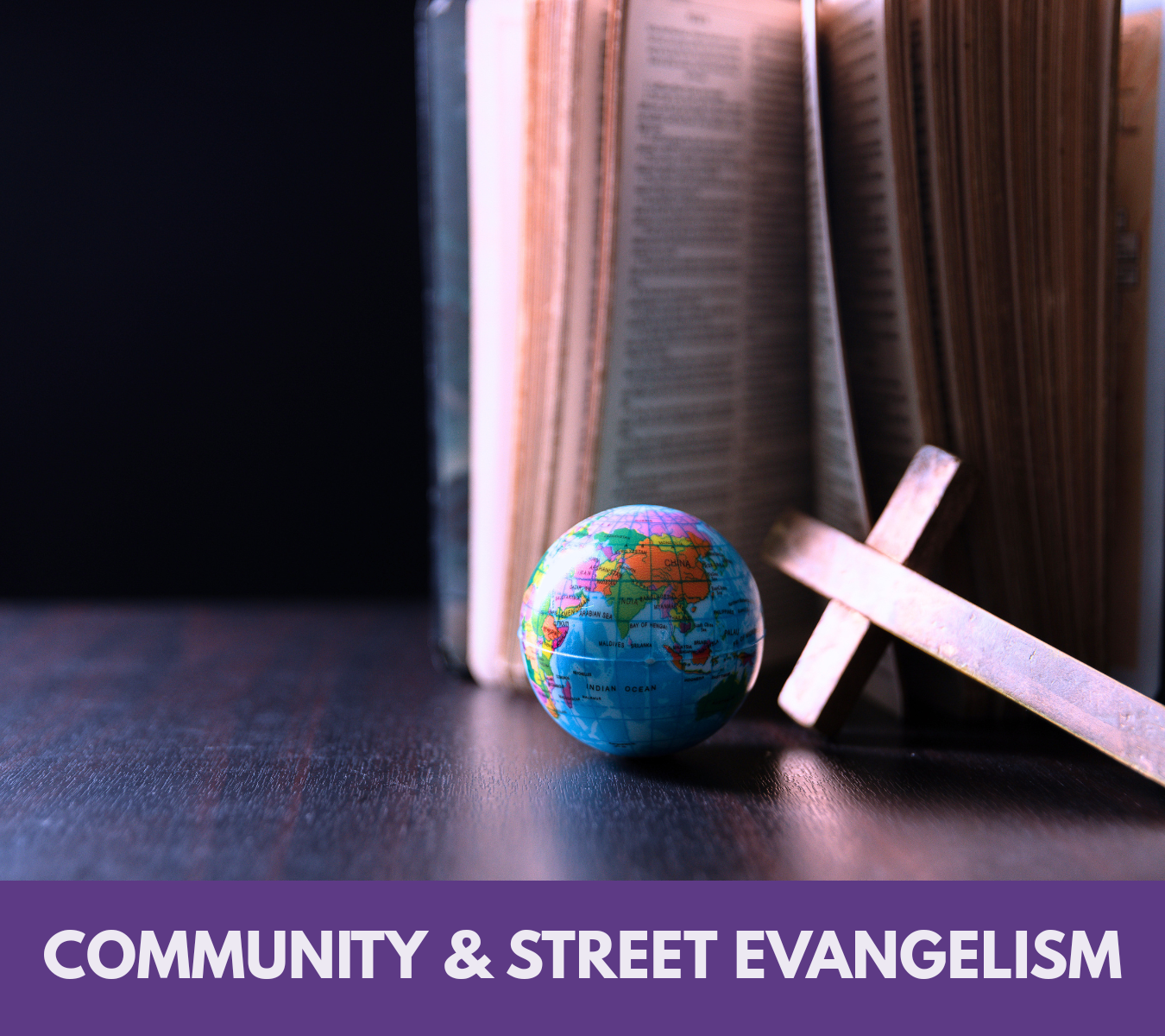A small globe, an open book, and a cross-shaped object on a dark surface with a dark background. The bottom caption reads 'Community & Street Evangelism.'