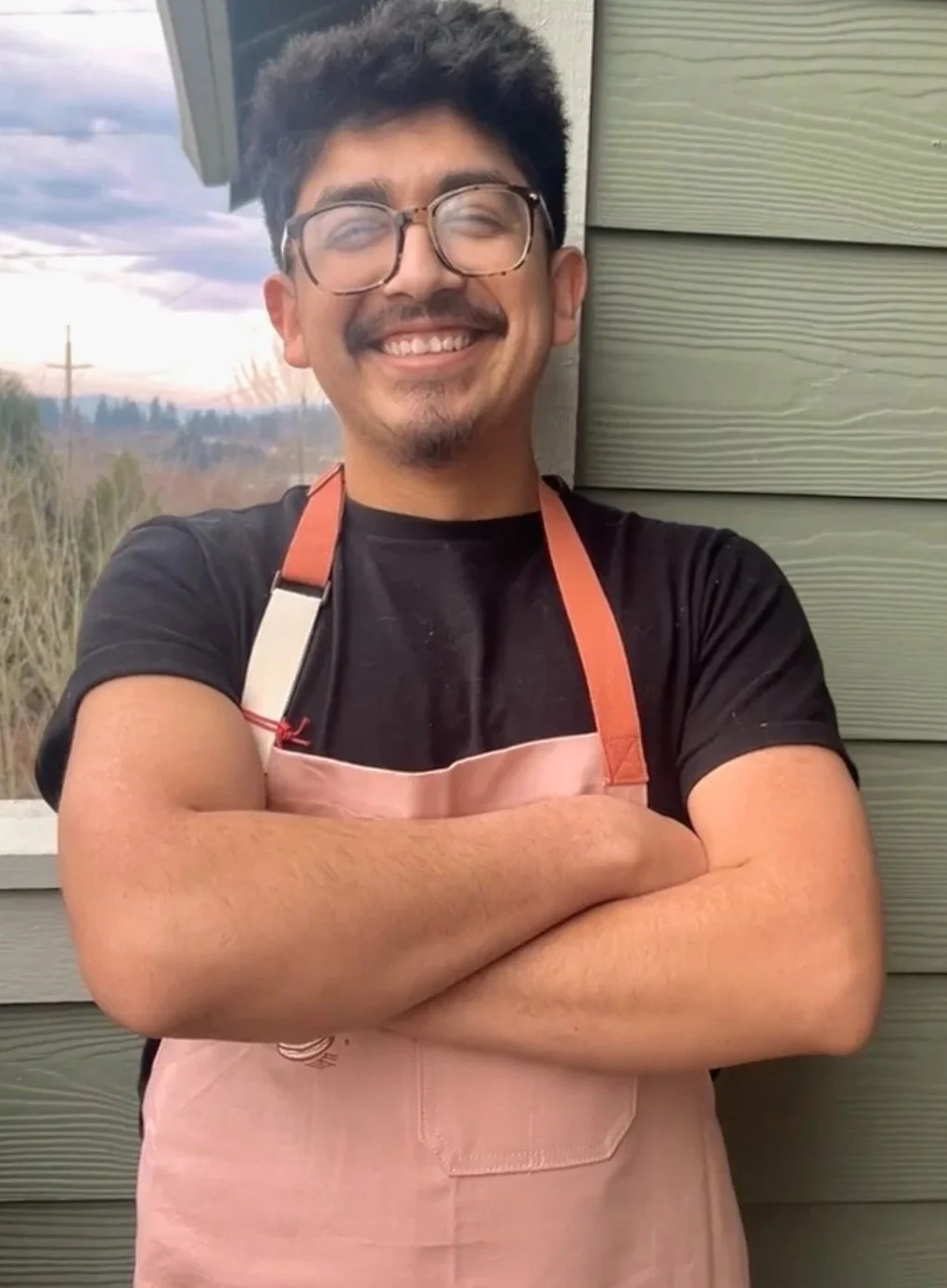 A young man with glasses, dark hair, and a beard, smiling with a crossed-arm pose, wearing a black T-shirt and a pink apron with orange straps, standing outdoors against a green wooden wall.