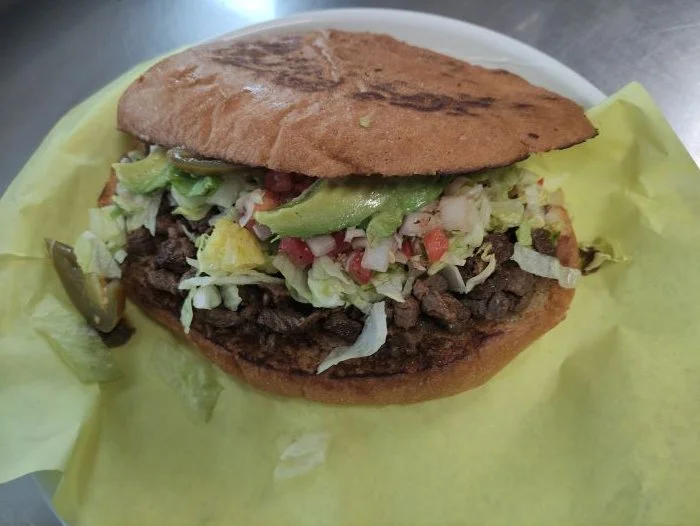 Close-up of a beef torta with chopped lettuce, tomatoes, and avocado in a toasted bread roll, served on yellow paper.