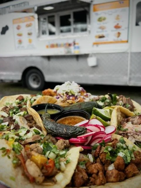 Close-up of a plate of tacos with grilled meats, pickled radishes, and sauces, with a food truck in the background.