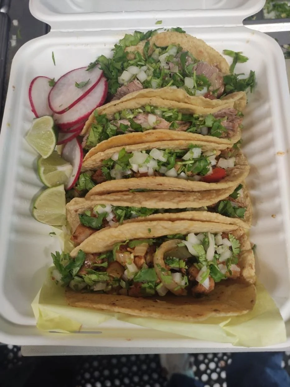 Six tacos filled with chopped meat, onions, cilantro, and vegetables, served with lime wedges and radish slices on the side.