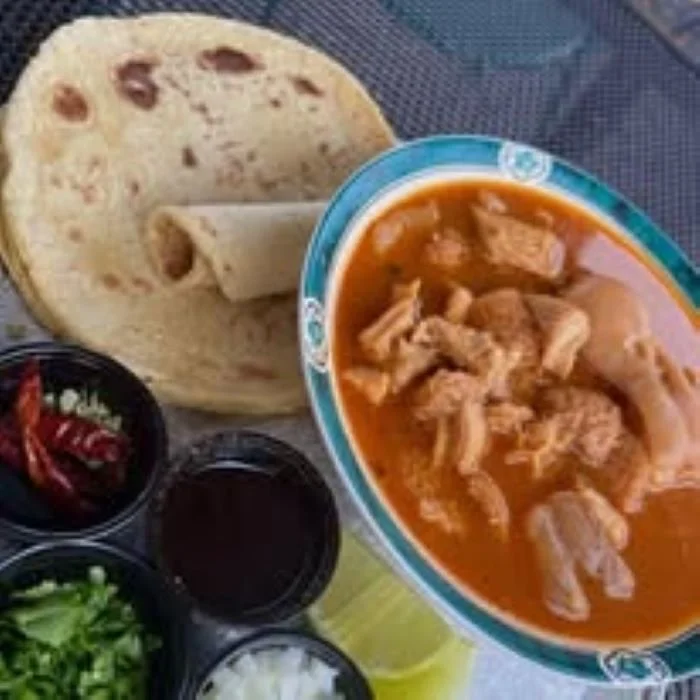 Bowl of Beef Tripe Menudo with a side of tortillas, chopped onions, cilantro, herbs, and dried red chili peppers nearby.