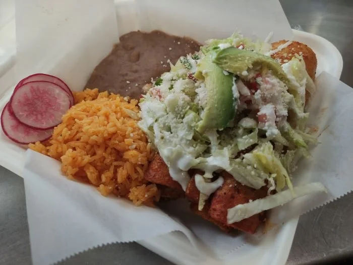Enchiladas with rice, refried beans, and tacos topped with lettuce, cheese, avocado, and sour cream, alongside sliced radishes
