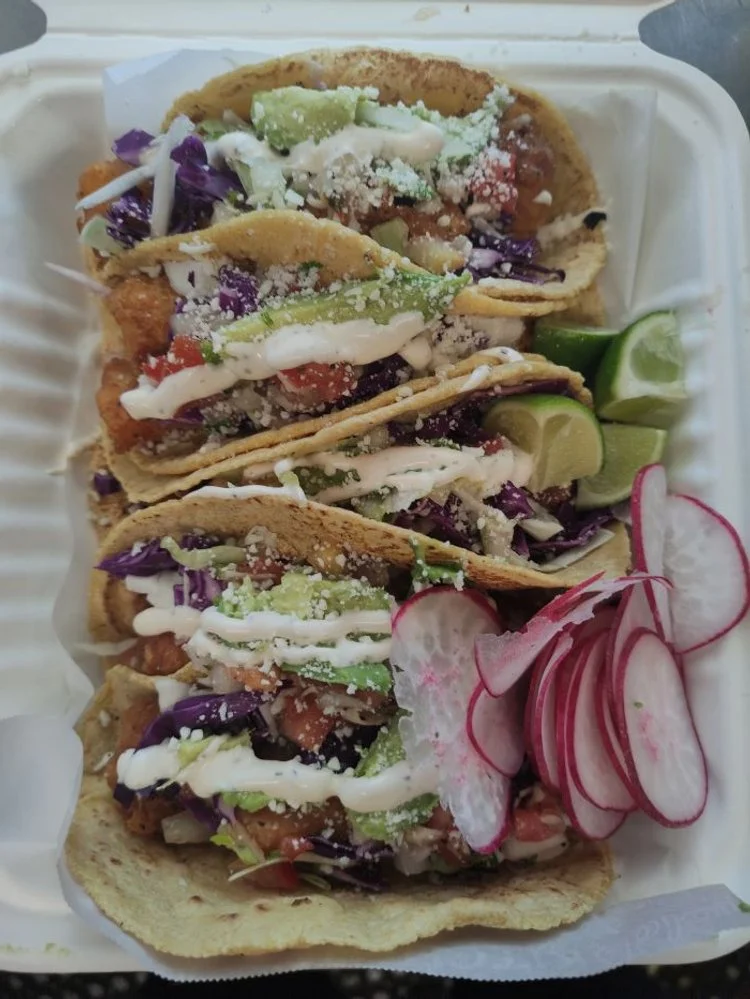 Three tacos filled with fried fish, shredded purple cabbage, chopped tomatoes, sliced cucumbers, and drizzled with white sauce, served with lime wedges and radish slices on the side.