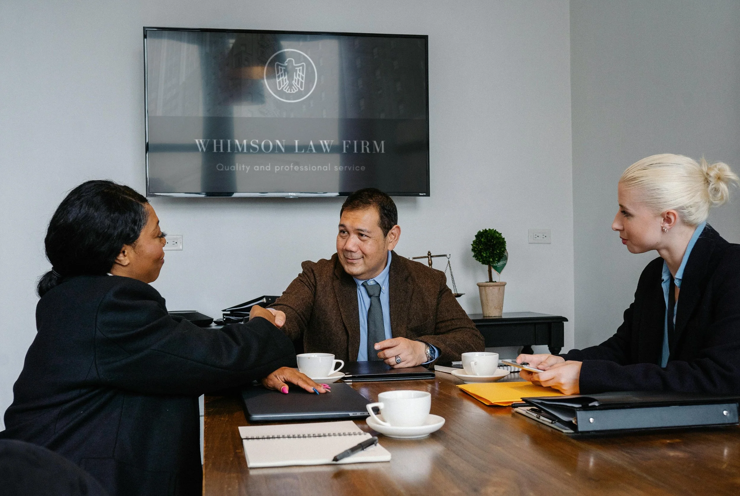 Two women and one man sitting at a conference table in a professional office, shaking hands. The woman on the left has dark hair and is wearing a black blazer. The man in the middle has short dark hair, a brown blazer, and a tie. The woman on the right has blonde hair tied back, wearing a black blazer. There are cups of coffee, notebooks, and folders on the table. A large monitor on the wall displays a logo for WHIMSON LAW FIRM, with the text 'Quality and professional service.'