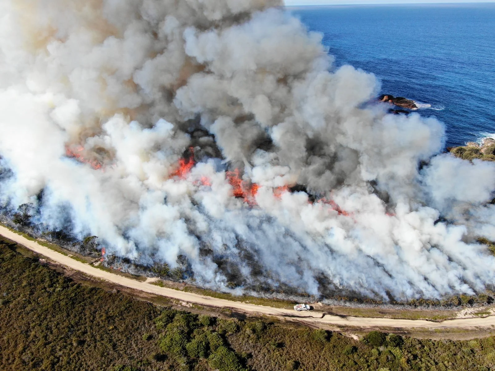 A planned hazard reduction burn near a coastal community and popular camping areas with thick smoke and flames, seen from above, with a dirt road and a vehicle nearby.