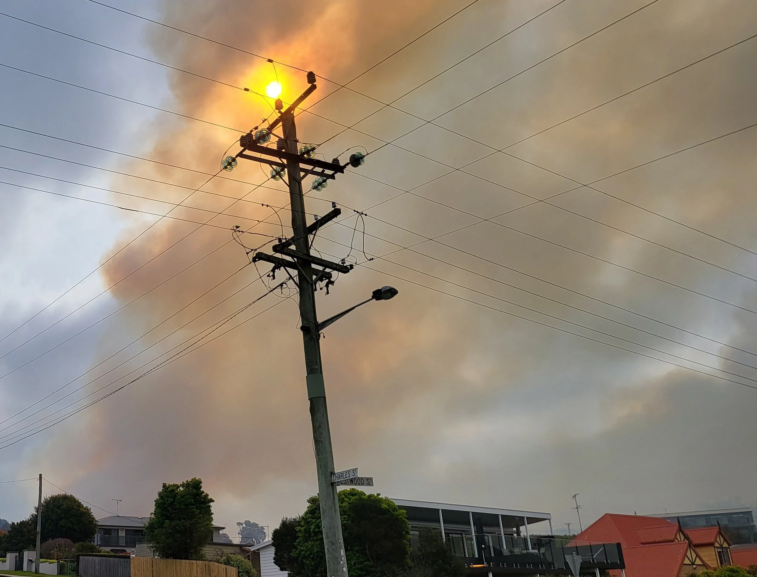 Telephone pole with power lines and streetlights, orange flames and smoke in the sky from a nearby fire, residential houses and trees below.