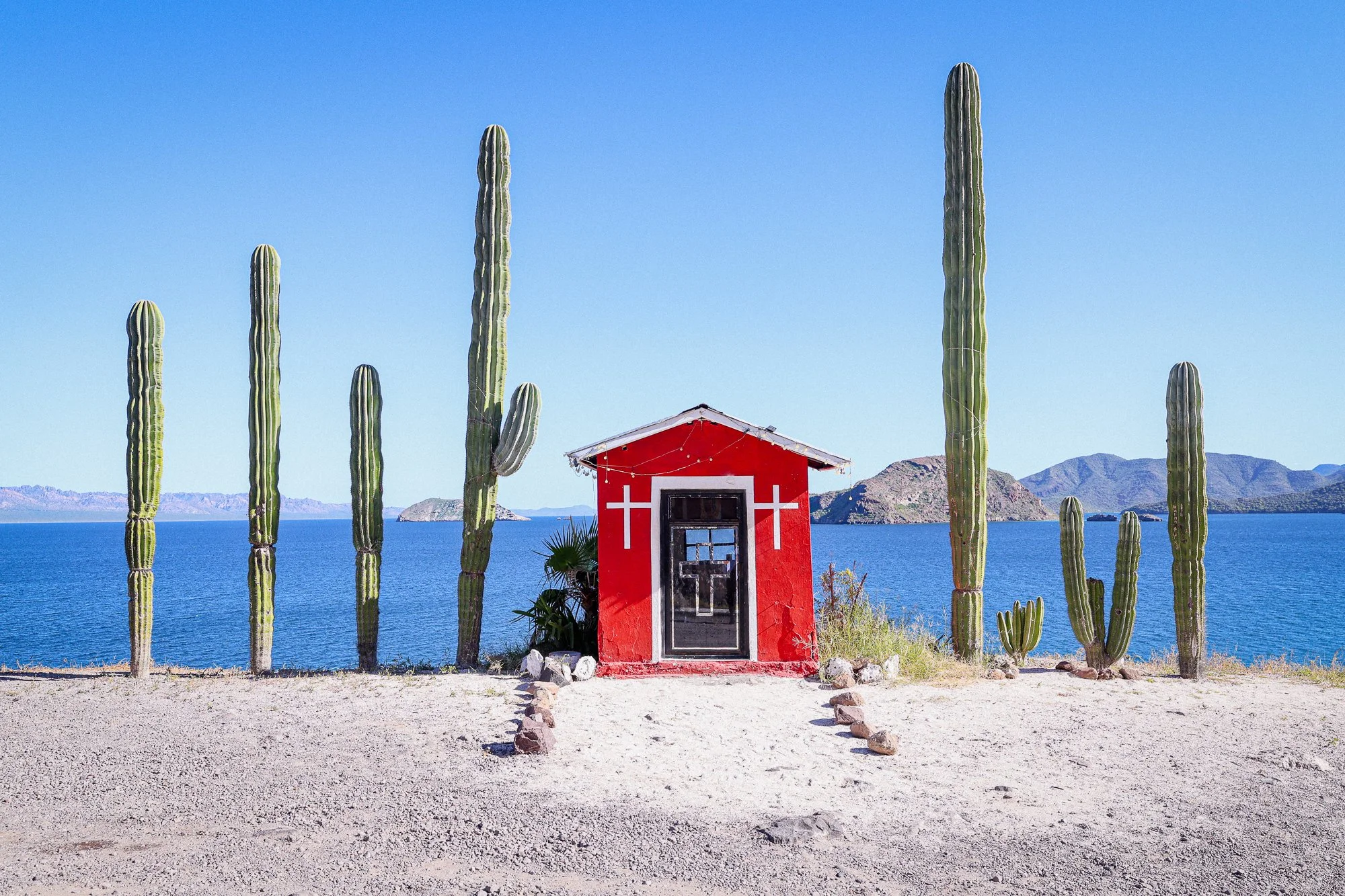 Una pequeña capilla roja con cruces blancas, rodeada de cactus, frente a un lago con montañas al fondo, en un paisaje árido y soleado.