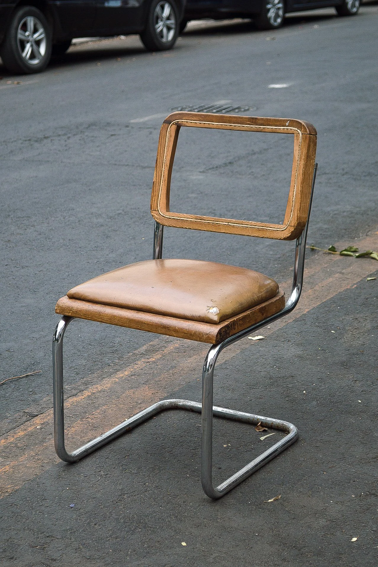 Silla de oficina antigua con estructura de metal, asiento de cuero desgastado y respaldo de madera con forma rectangular, ubicada en una calle con autos estacionados.