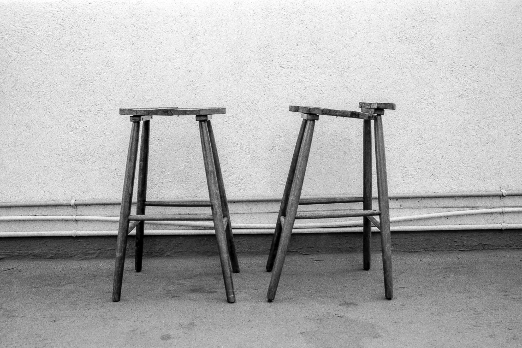 Dos sillas de madera sin asientos, una de ellas con una pata rota, colocadas contra una pared blanca y un piso de concreto