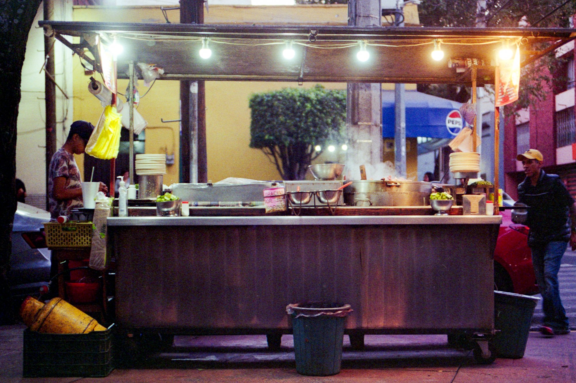 Un puesto de comida callejera en la noche, con luces colgantes y vapores saliendo de las ollas, dos personas atendiendo, en una ciudad con edificios coloridos y árboles.