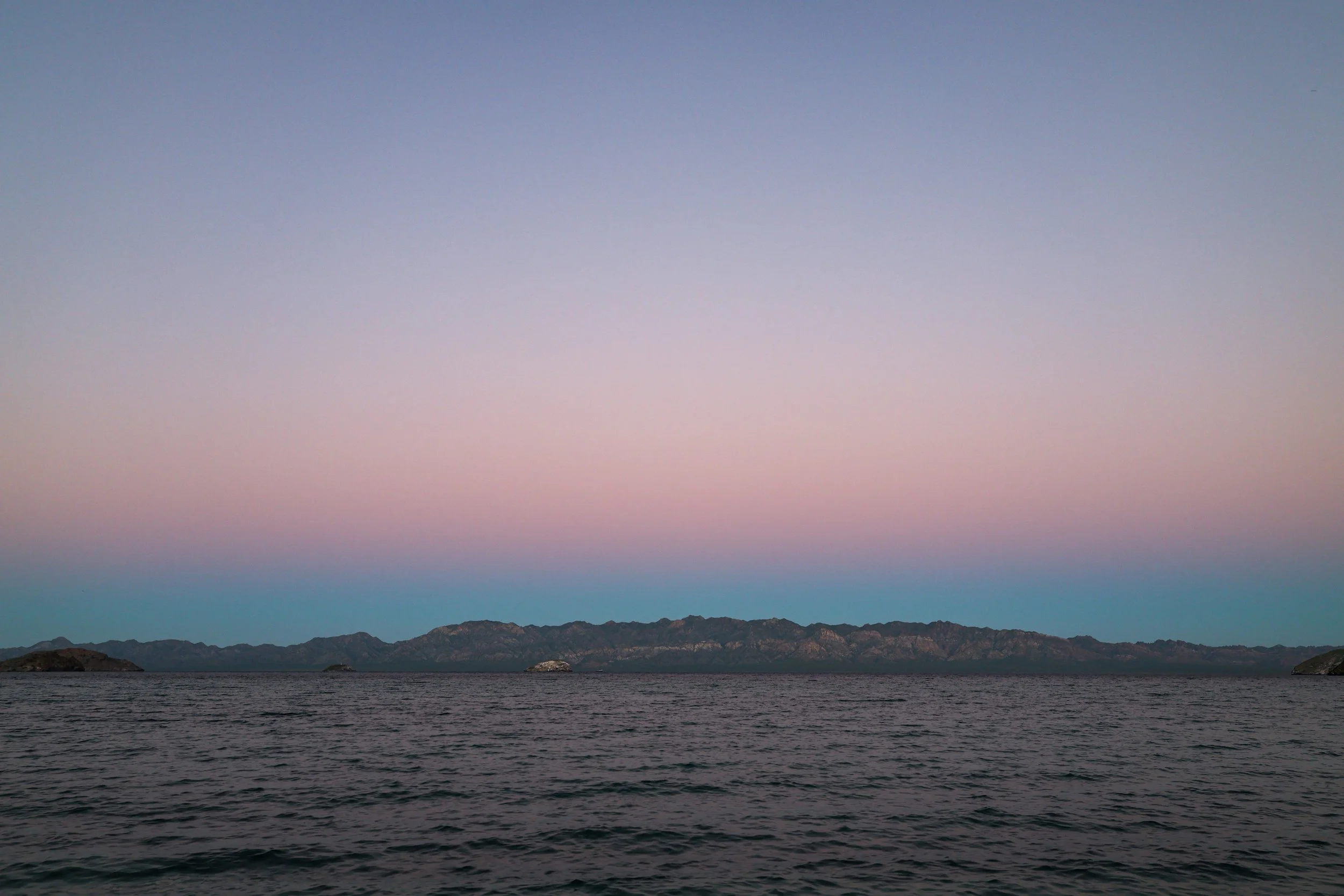 Paisaje de mar con olas suaves, montañas en la distancia y un cielo al atardecer con tonos pastel de azul, rosa y púrpura.