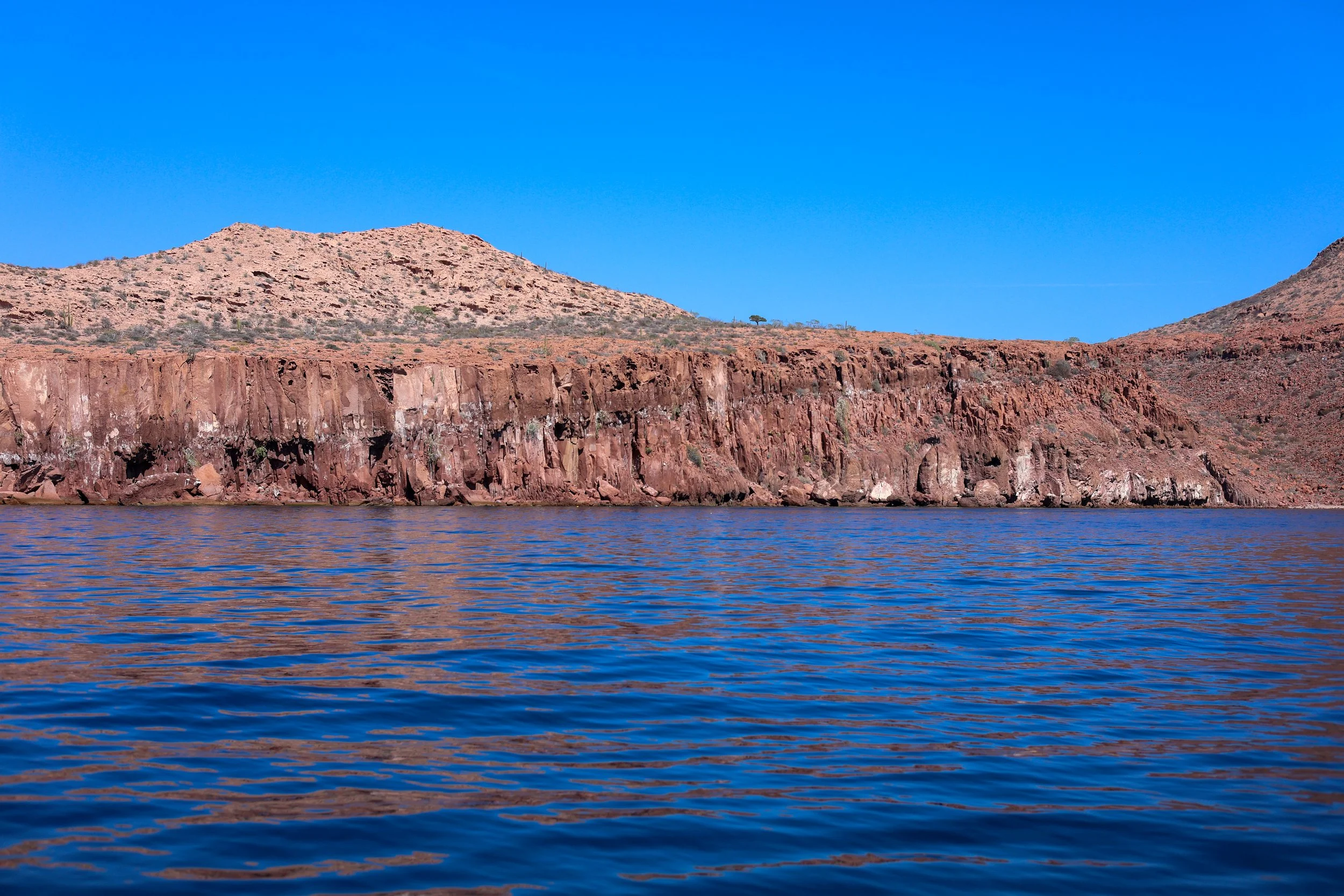 Paisaje de un cuerpo de agua con rocas y montañas áridas de color marrón y beige bajo un cielo azul claro.