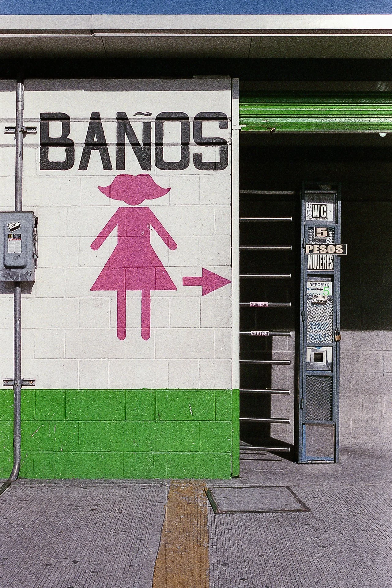 Señal de un baño para mujeres con una figura femenina en color rosa y una flecha apuntando a la derecha, junto a un cartel que dice 'Baños Mujeres' y una cabina de pago.