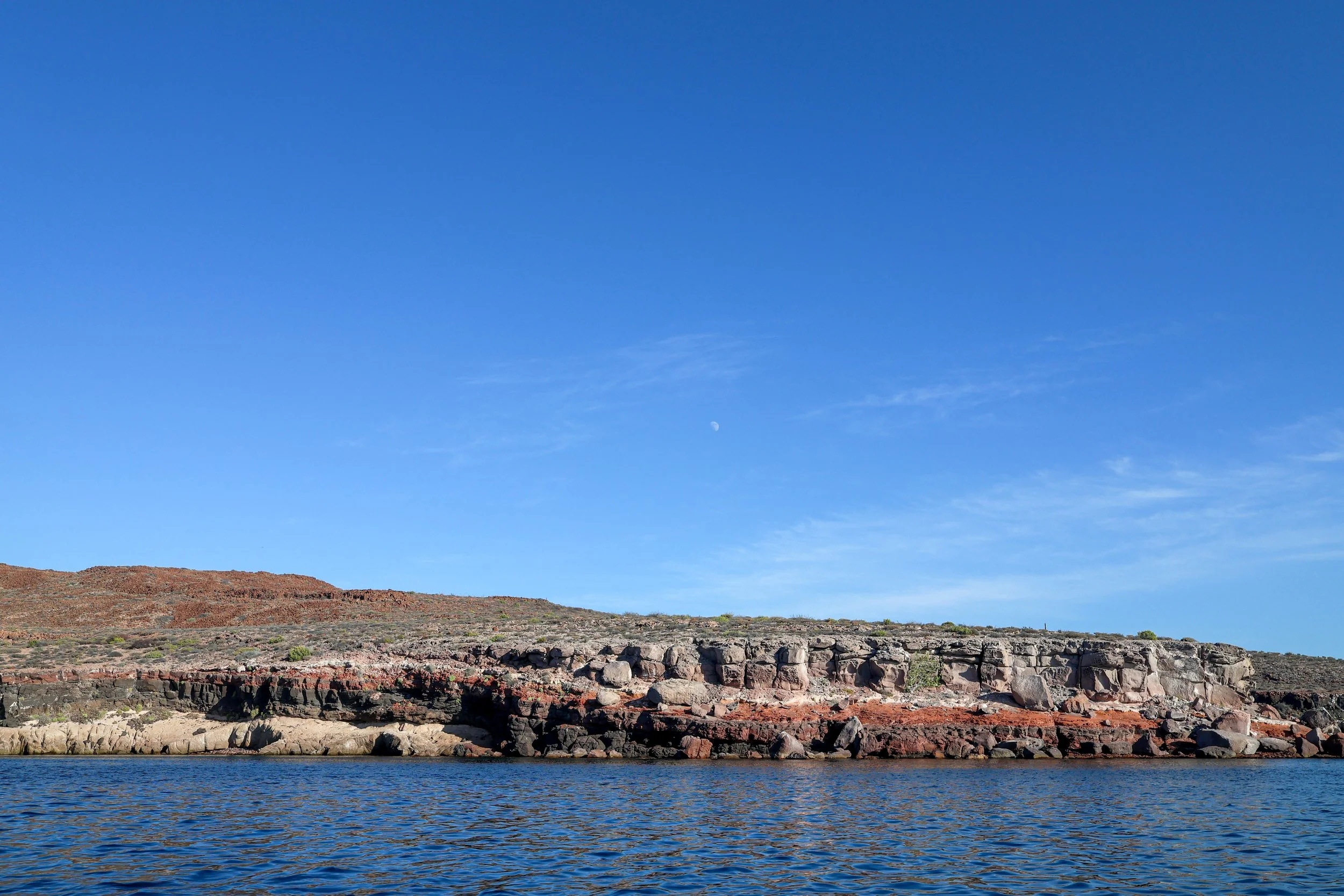 Paisaje con agua en primer plano, rocas y tierra en el fondo, cielo despejado con algunas nubes y luna visible.
