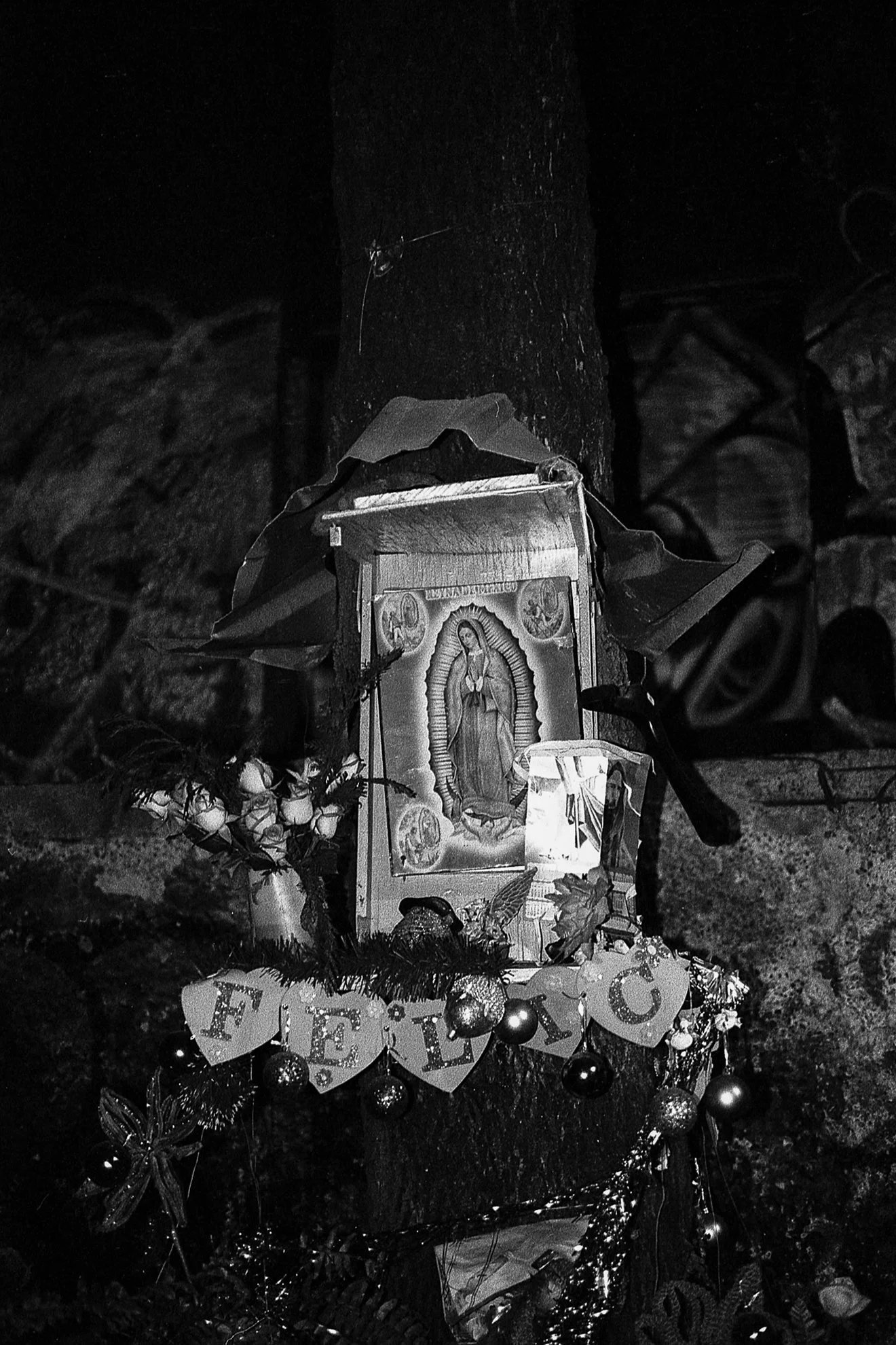 Una ofrenda con la imagen de la Virgen de Guadalupe, decorada con flores, papel decorativo y adornos navideños, colocada en un árbol o poste en un contexto nocturno.