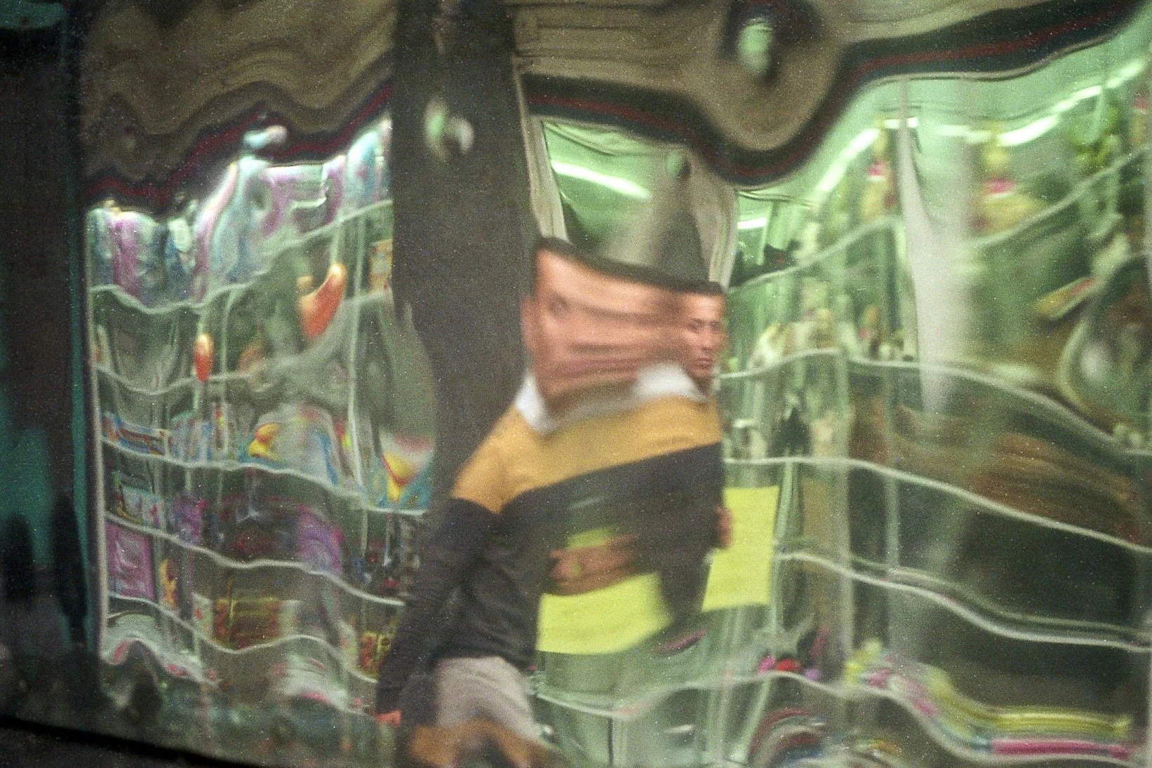 Reflejo de una persona con una camiseta amarilla y negra, en una superficie metálica que parece un espejo deformado.