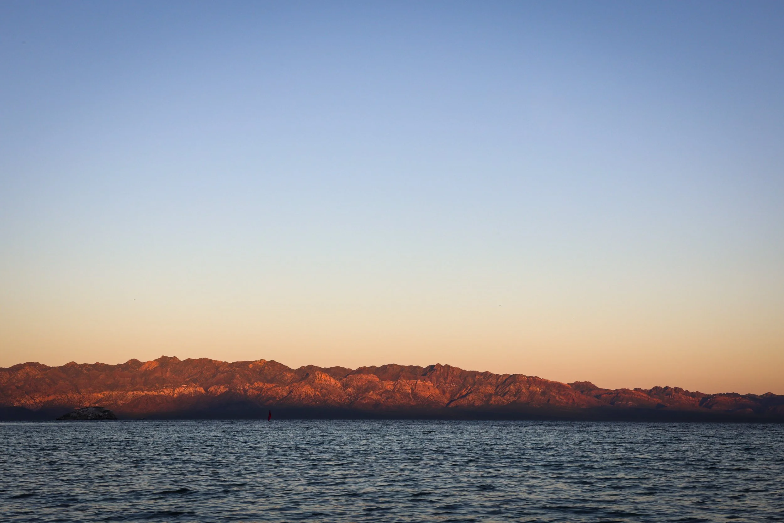 Monte con picos rojos al atardecer, agua en primer plano bajo un cielo despejado