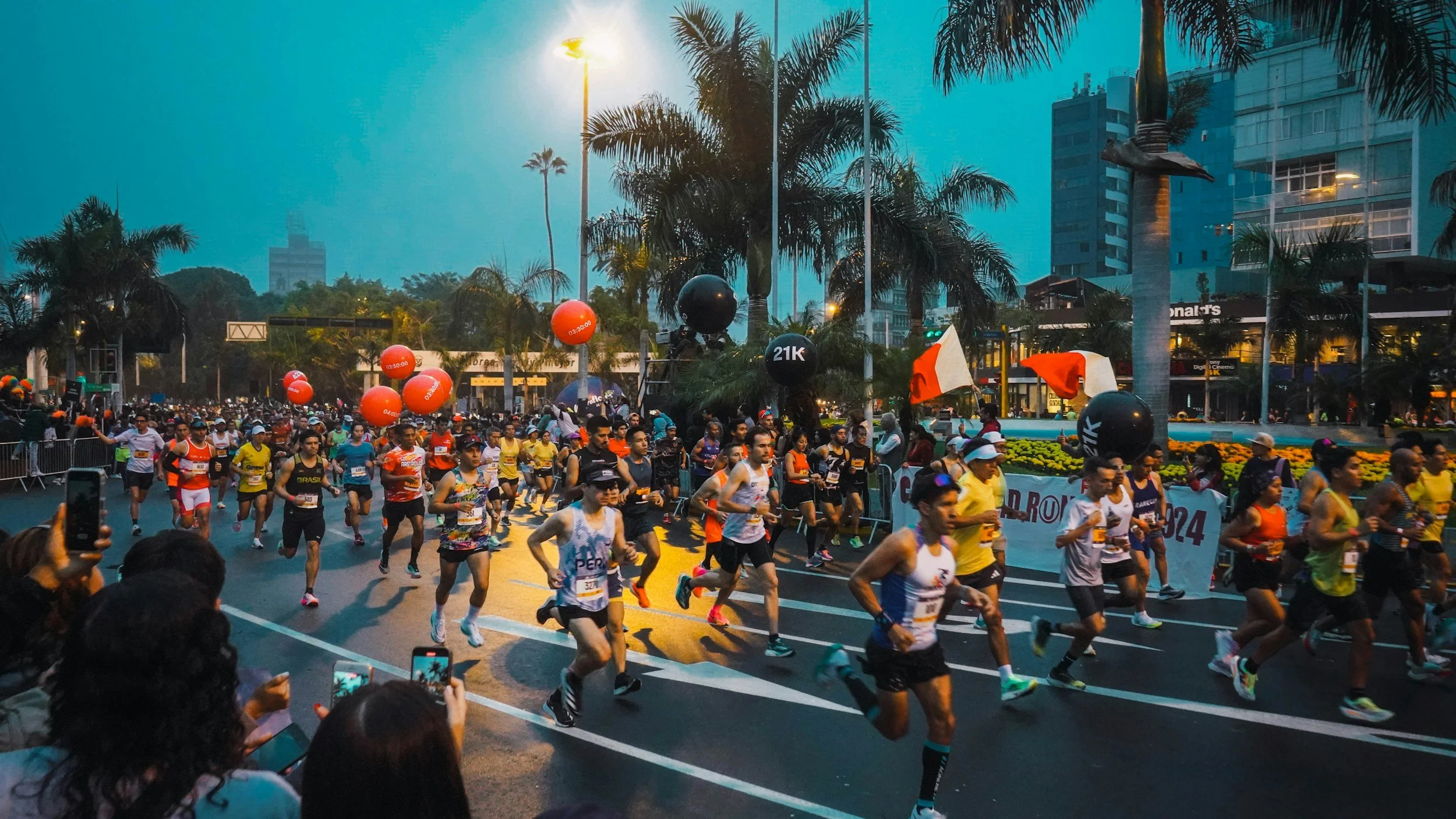 A large group of runners participating in a marathon or race on a city street is shown. The scene takes place during twilight or early evening, with tall buildings and palm trees in the background. Spectators are visible along the sides, some taking photos. Balloons and flags are part of the event decorations.