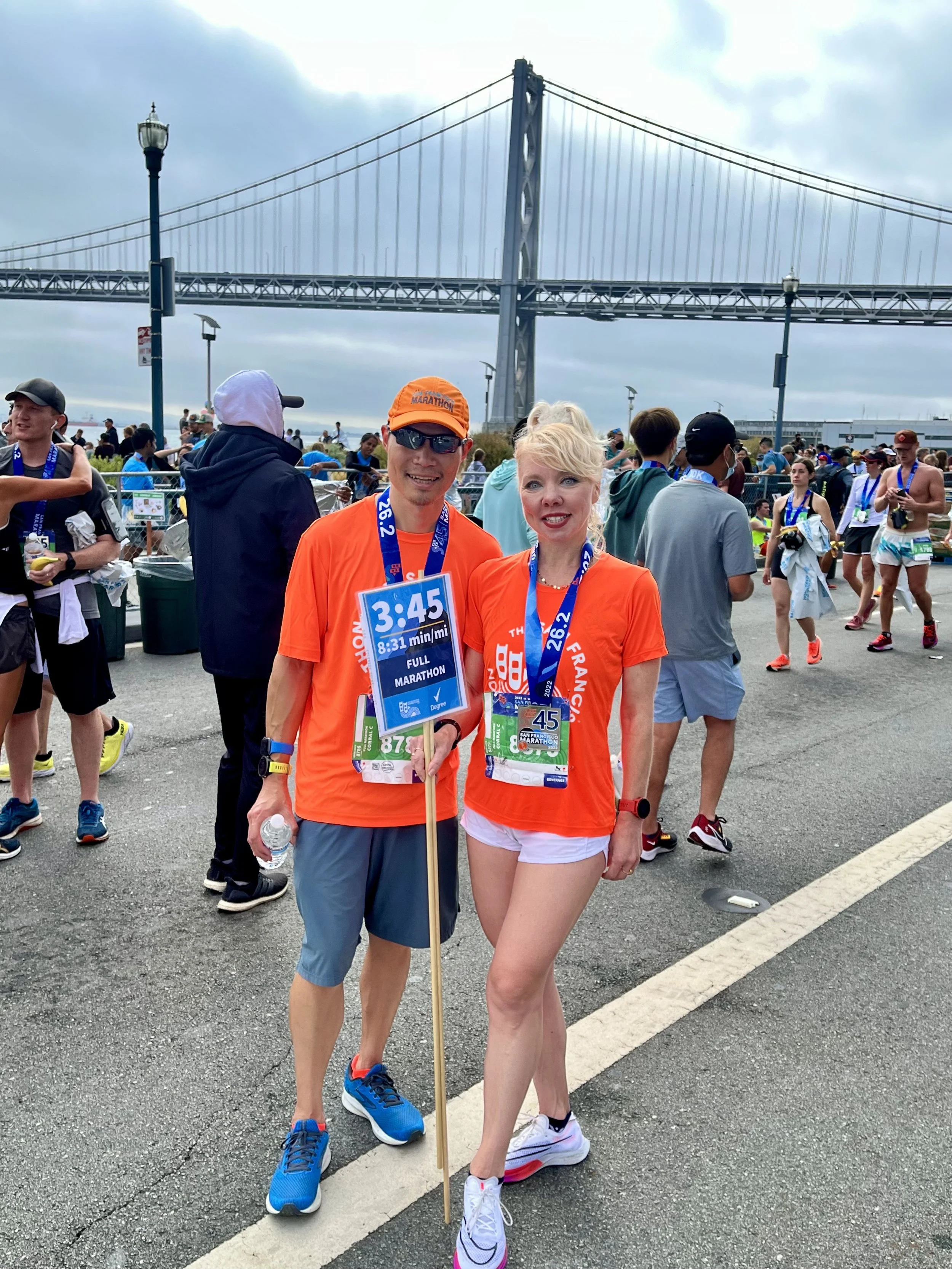 Two marathon runners, a man and a woman, standing together at a race finish area with the San Francisco-Oakland Bay Bridge in the background. Both are wearing bright orange shirts and smiling.