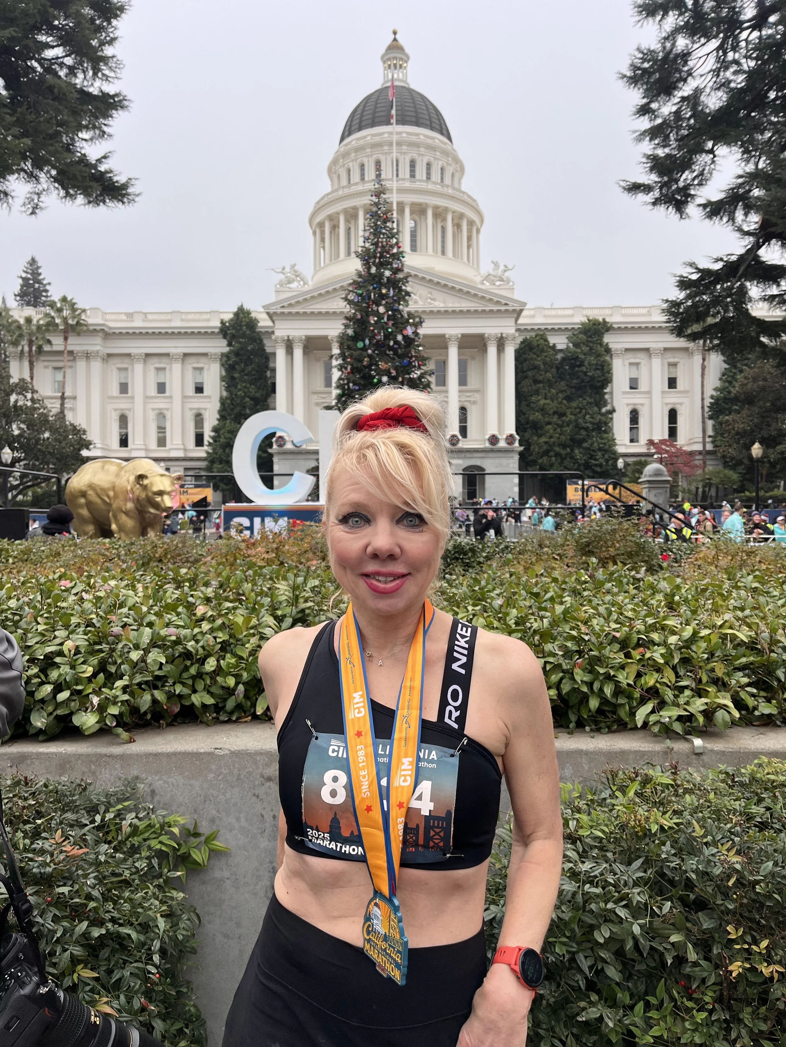 A woman standing in front of the California State Capitol building during a marathon event, wearing medals and race bibs, with a decorated Christmas tree in the background.