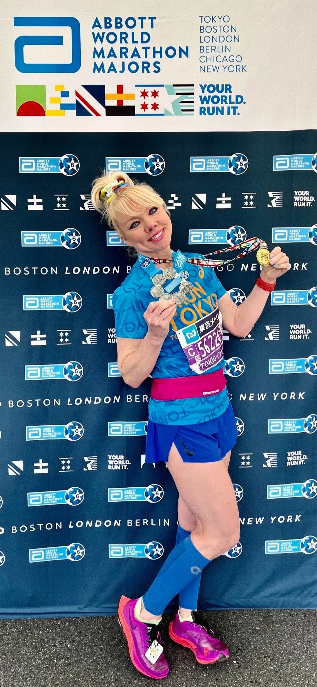 A woman in running attire holding medals after finishing all 7 Abbott marathons, standing in front of a backdrop with marathon logos, celebrating her achievement.
