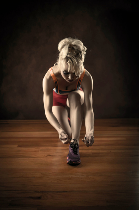 Young woman tying her running shoe on a wooden floor, wearing athletic clothing, in a dark studio setting.