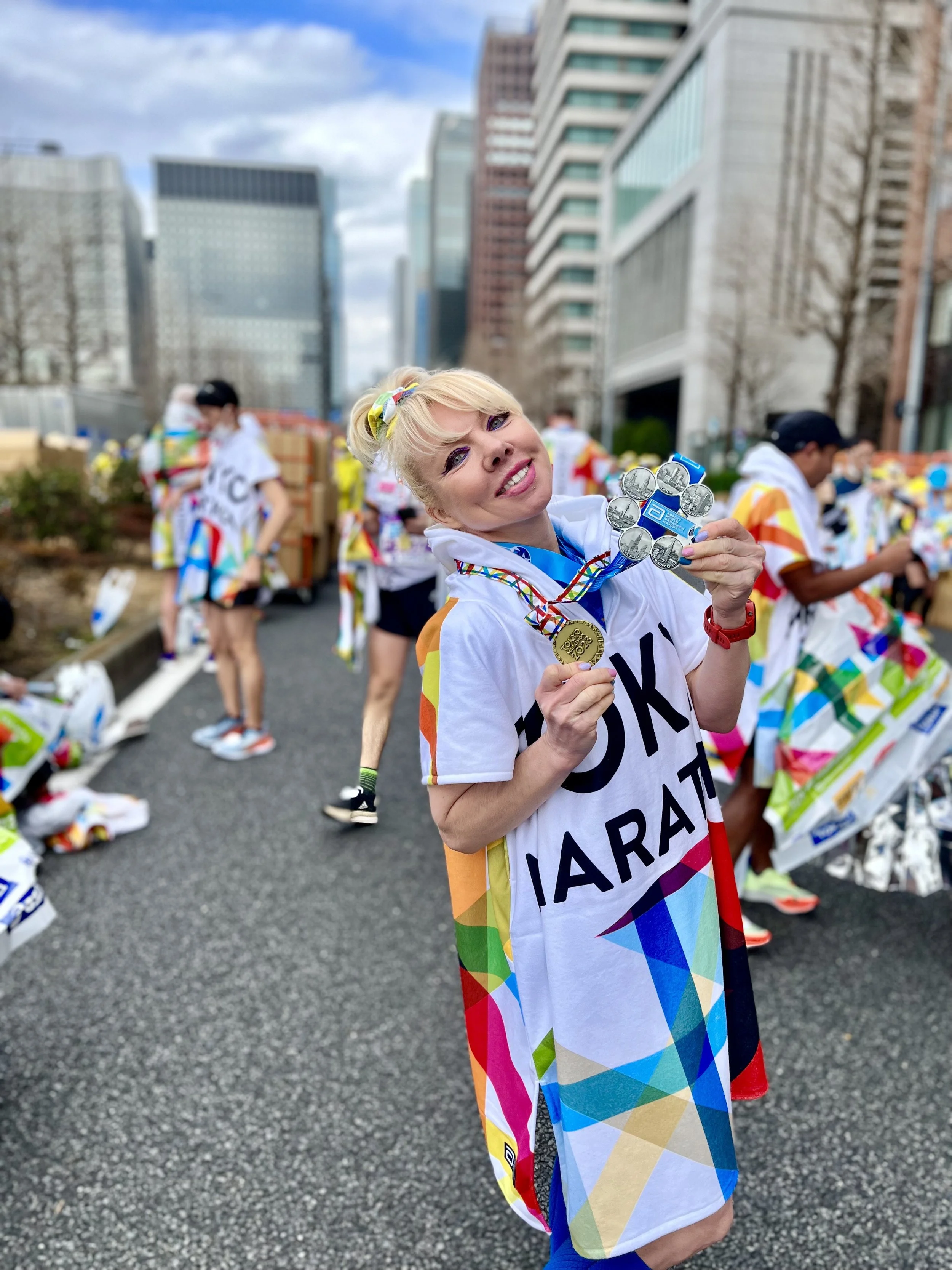 A smiling woman with blonde hair and colorful makeup, holding medals at a marathon event in an urban area, wearing a T-shirt that says 'UKI MARATHON' with a rainbow-colored towel draped over her shoulders, with other runners and buildings in the background.