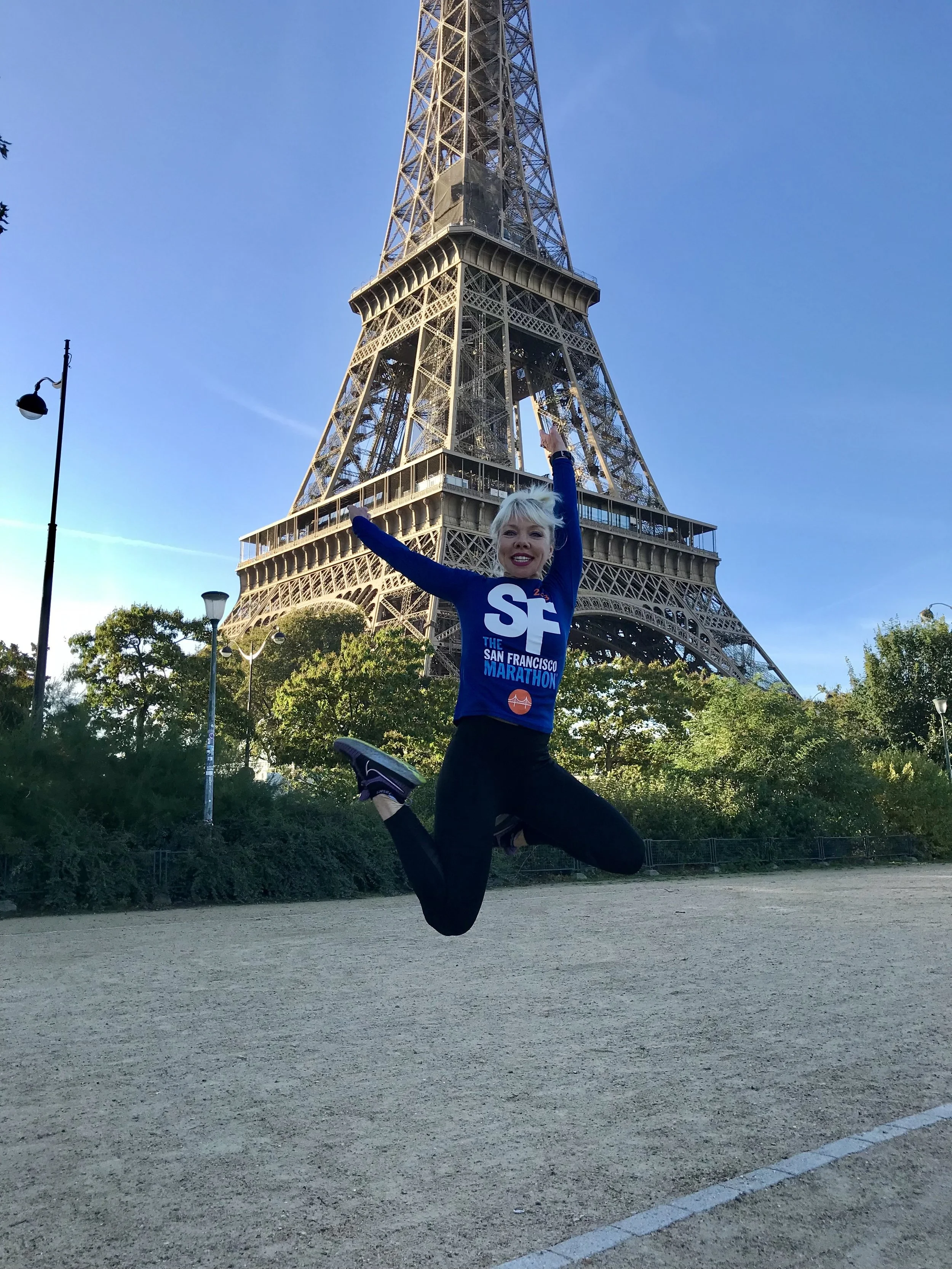 A woman with blonde hair jumps in the air with arms outstretched in front of the Eiffel Tower in Paris, wearing a blue shirt for the San Francisco Marathon, on a clear day with a blue sky and trees in the background.