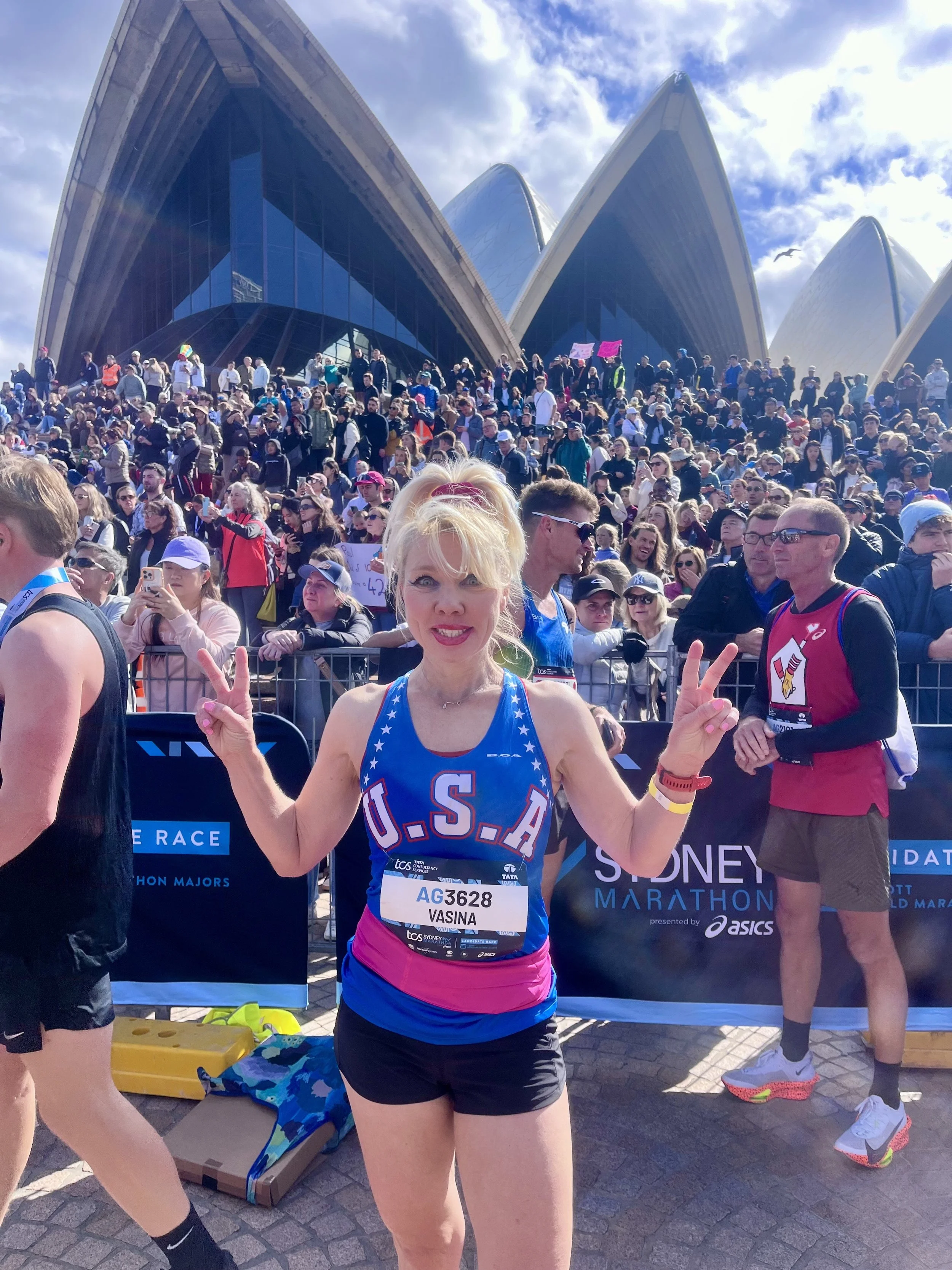 Female marathon runner at the Sydney Marathon, wearing an USA tank top, making peace signs with both hands, with the Sydney Opera House and a large crowd of spectators in the background.