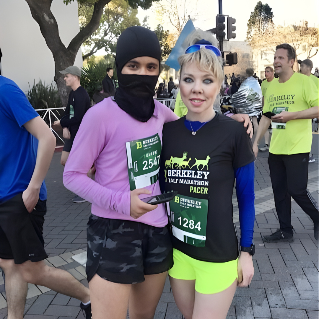Two women in athletic wear standing together at a marathon event, one with a pacer bib, holding a phone, and the other wearing a Berkeley marathon shirt with running shorts, in an outdoor setting with other runners and trees in the background.