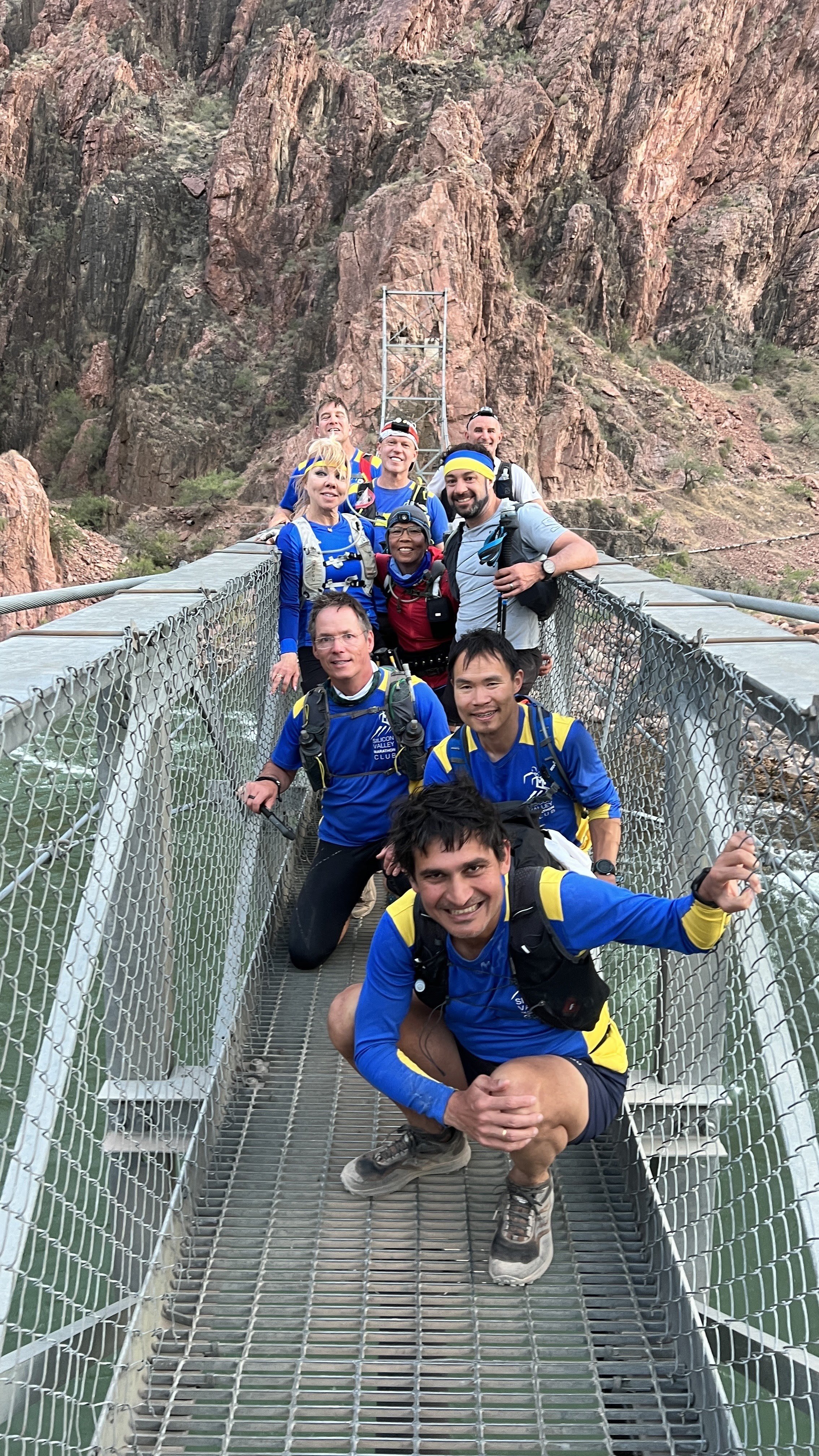 Group of people on a suspension bridge in a rocky, mountainous area, wearing hiking gear and smiling at the camera.