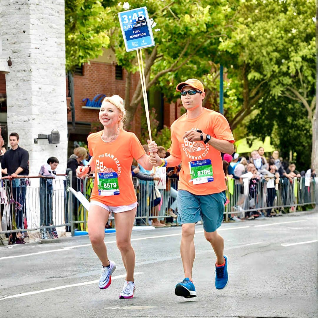 A woman and a man running in a marathon, holding a blue sign indicating they have completed 20 miles in 3 hours and 45 minutes, with spectators watching from the sidelines.