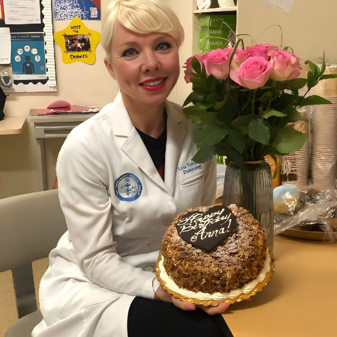 A woman in a white nurse coat holding a birthday cake with a message on a chocolate plaque that says "Happy Birthday Anna". She is sitting at a table with a vase of pink roses beside her.