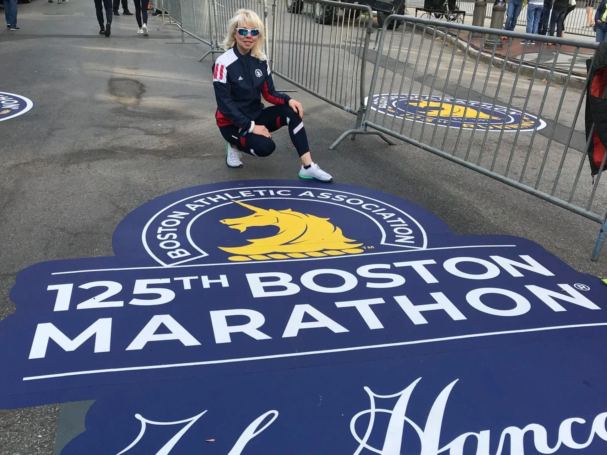 A woman kneeling next to a large sign for the 125th Boston Marathon, featuring the Boston Athletic Association logo and a yellow unicorn.