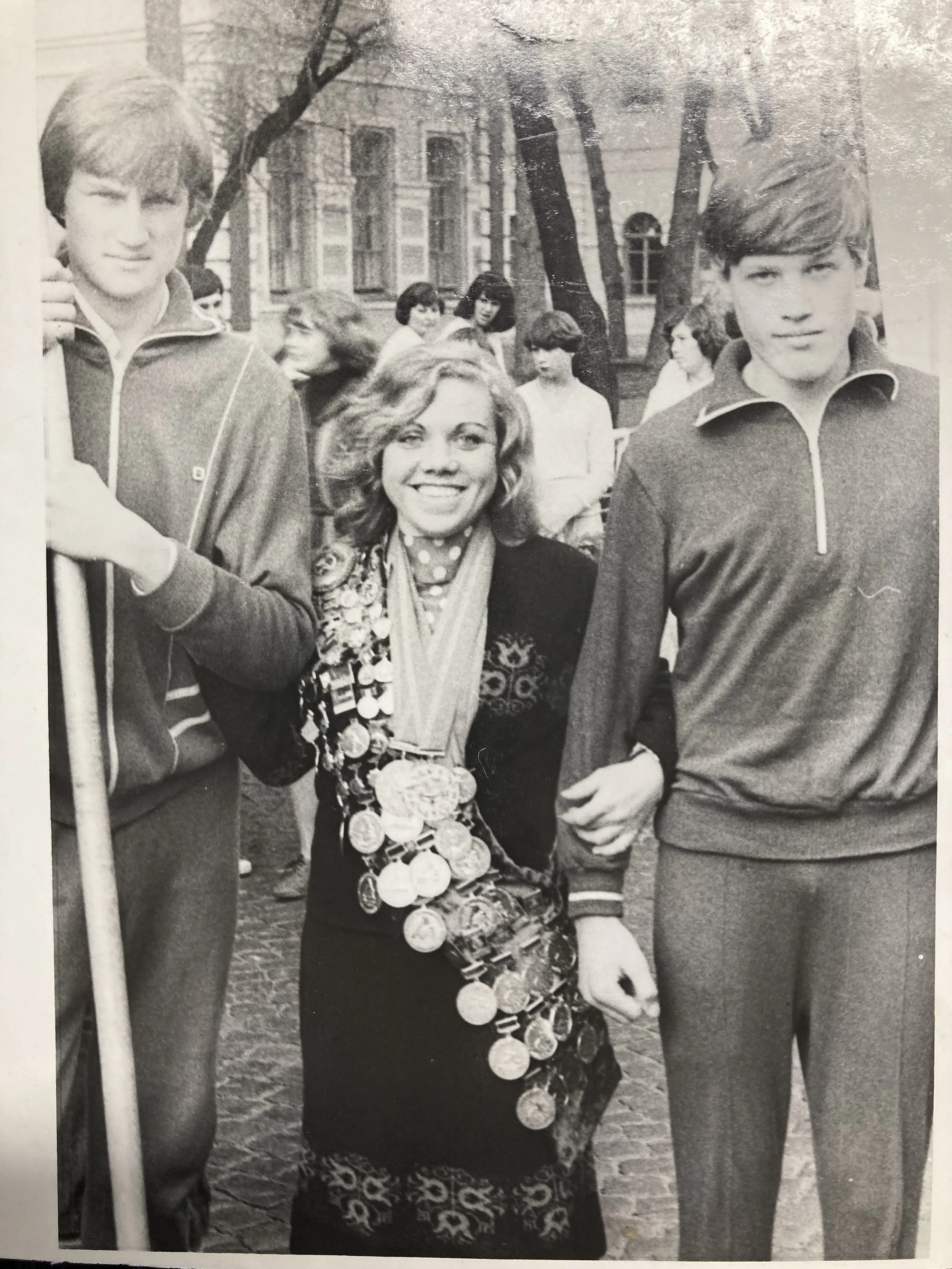 Black and white photo of a woman with a lot of medals around her neck, standing outdoors with two teenage boys, holding hands with one of them. The woman is smiling, and the boys are looking at the camera. Several people are in the background.