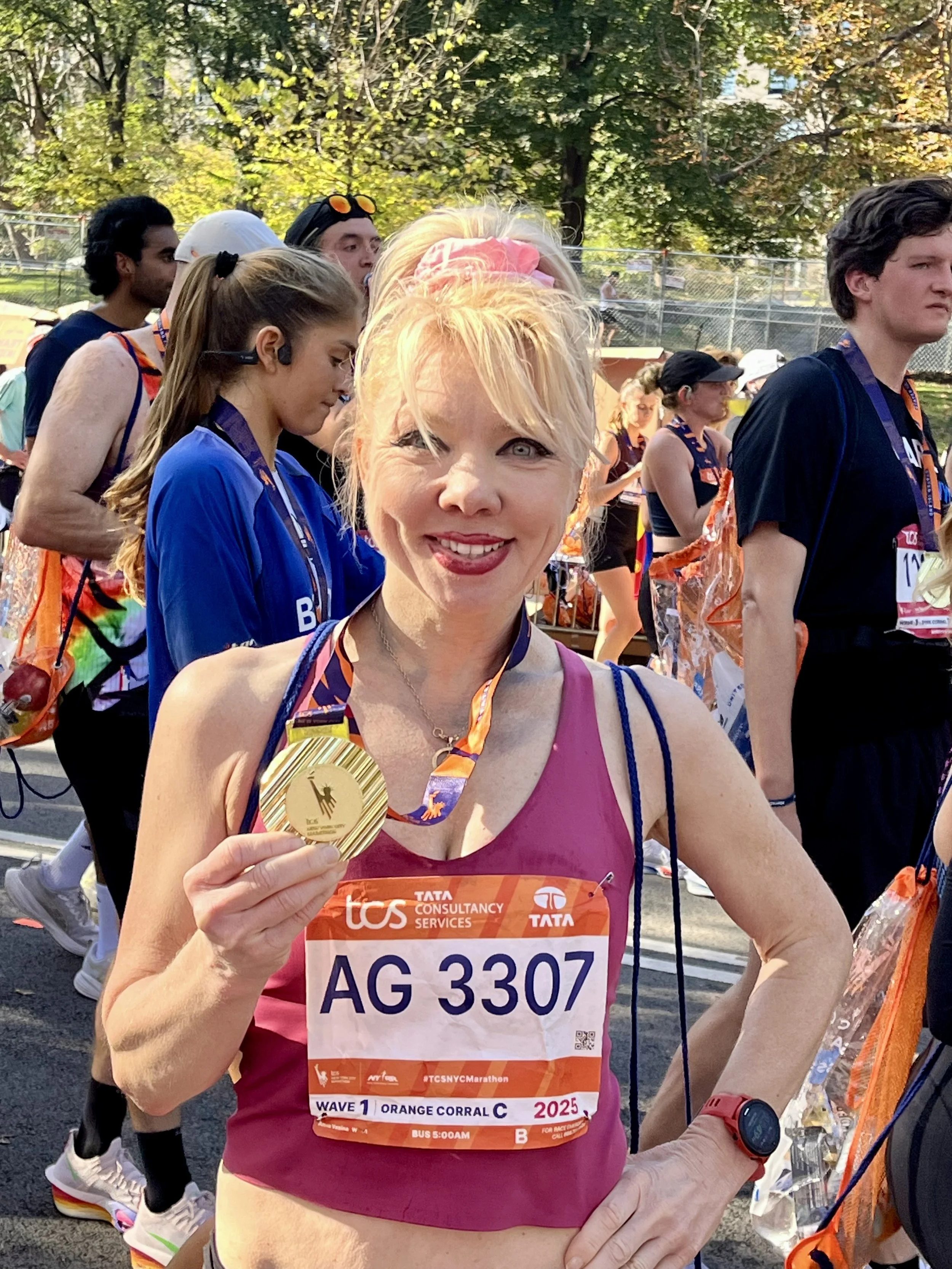 A woman smiling and holding a gold medal at a marathon event, wearing a pink tank top with a race bib, surrounded by other runners outdoors.