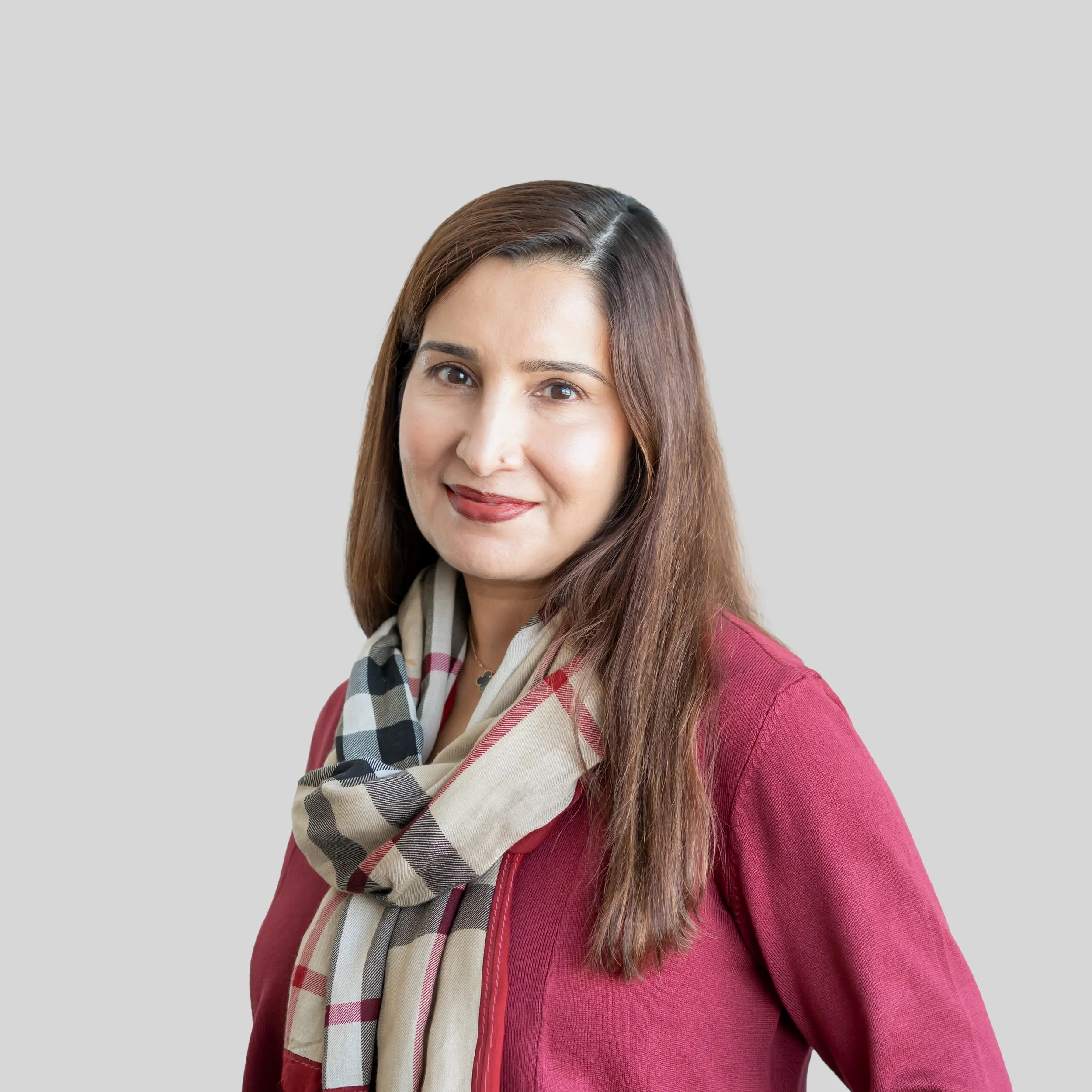 Dr. Tayyab, with long brown hair, wearing a red jacket and a beige checkered scarf, smiling.
