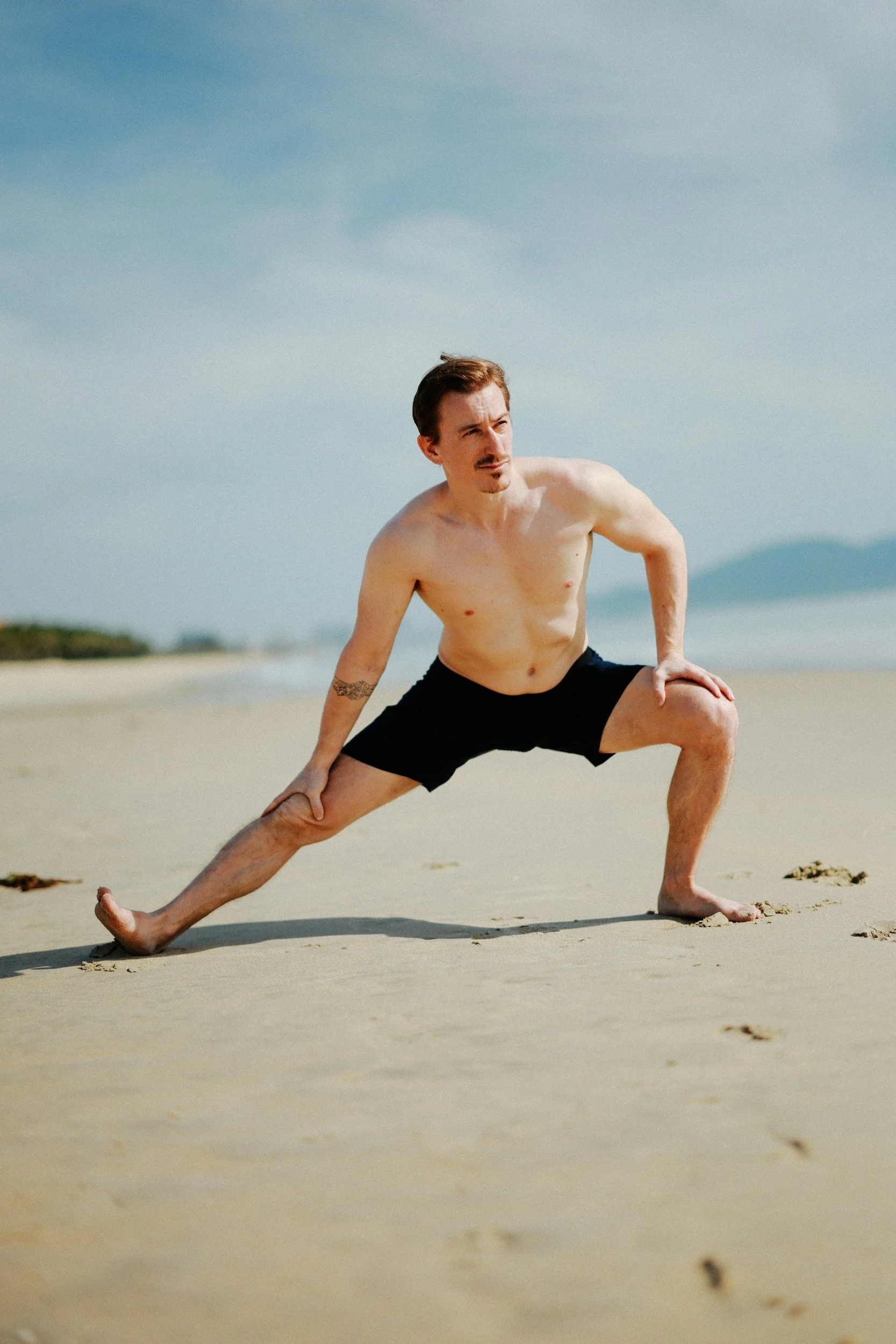 A man in black shorts performs a yoga pose on a sandy beach, with the ocean and a cloudy sky in the background.