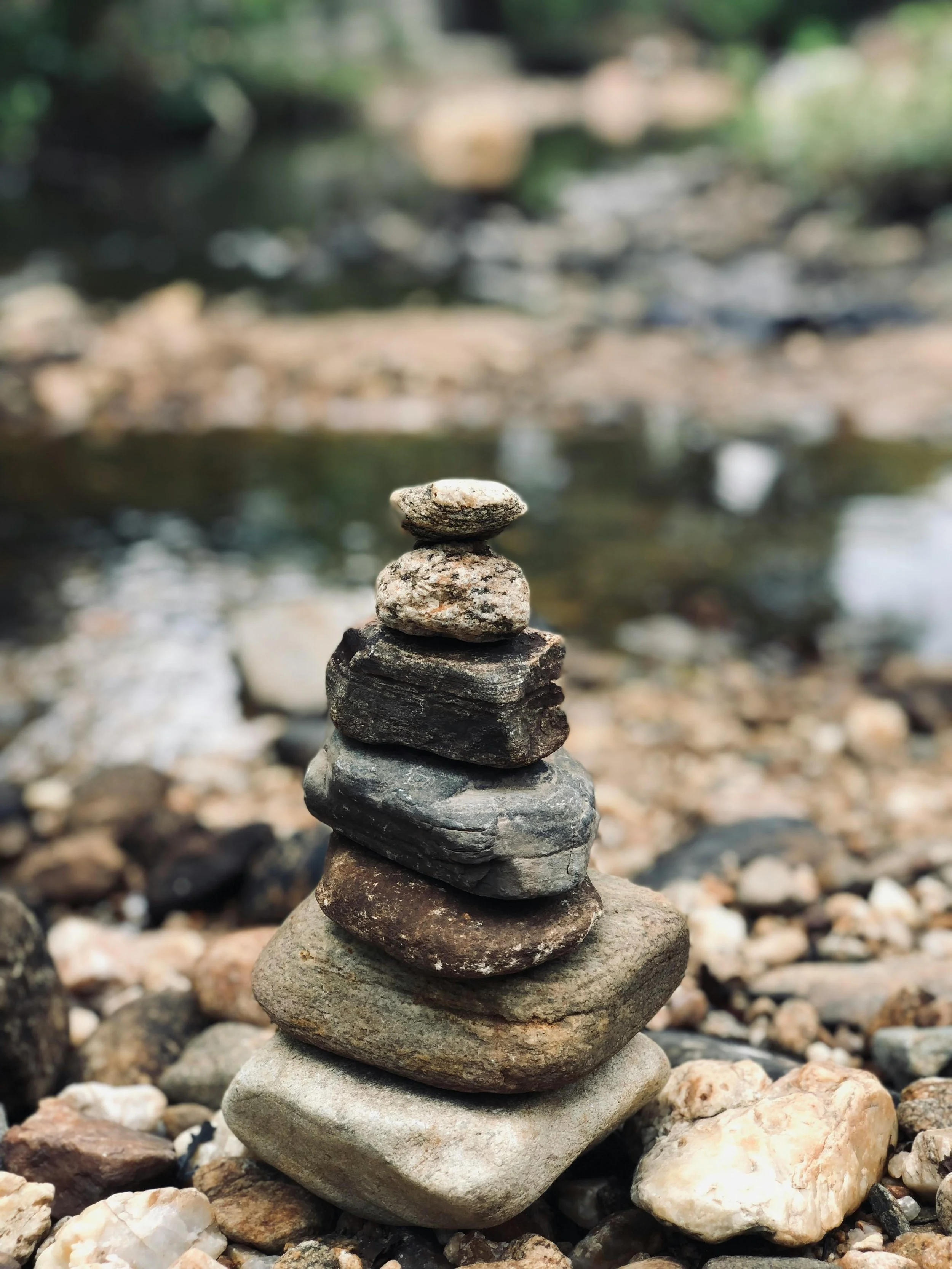 Stacked rocks on a rocky shoreline by a river with blurred trees in the background.