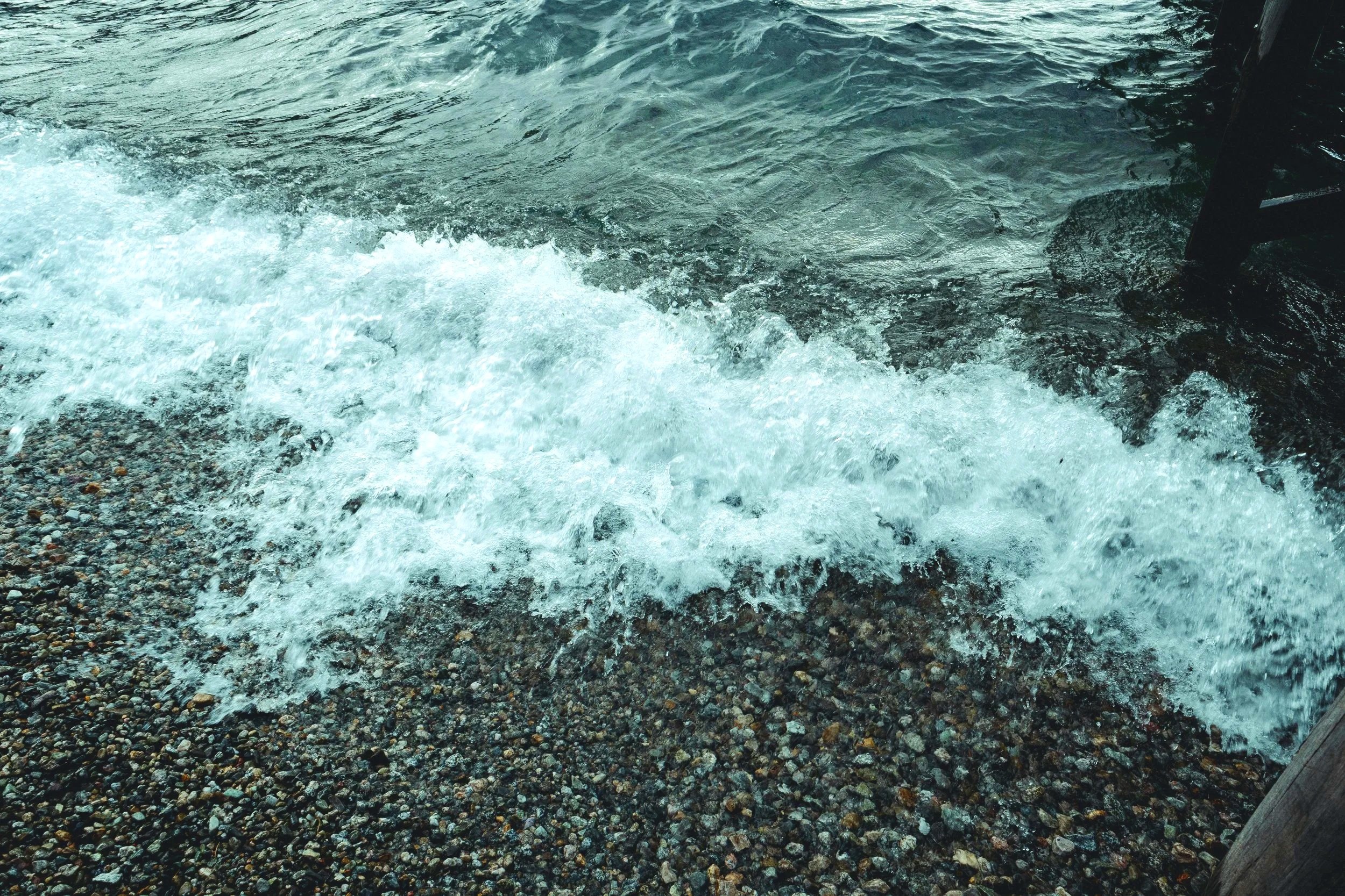 Waves crashing onto a rocky shoreline with dark water and a wooden structure at the edge.