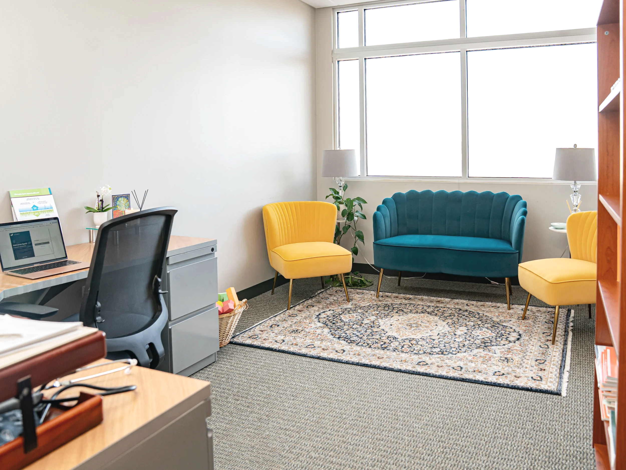 An office with a desk on the left, a black office chair, a laptop, and decorative items; on the right, a seating area with a teal velvet sofa, two yellow chairs, two white lamps, a potted plant, and a patterned rug in front of a large window.