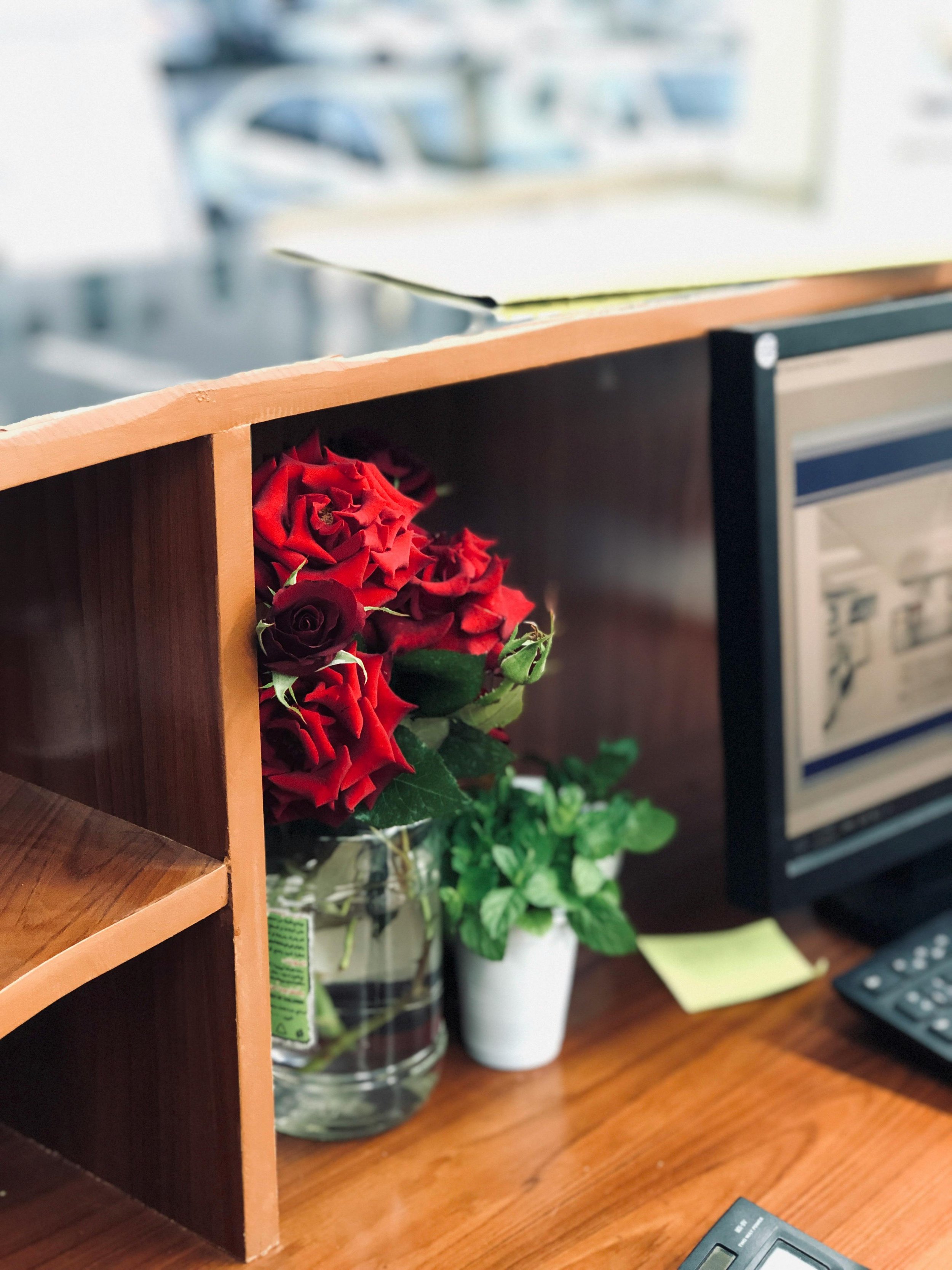 A bouquet of red roses and a vase of green leaves on a wooden desk, next to a computer monitor and remote controls.