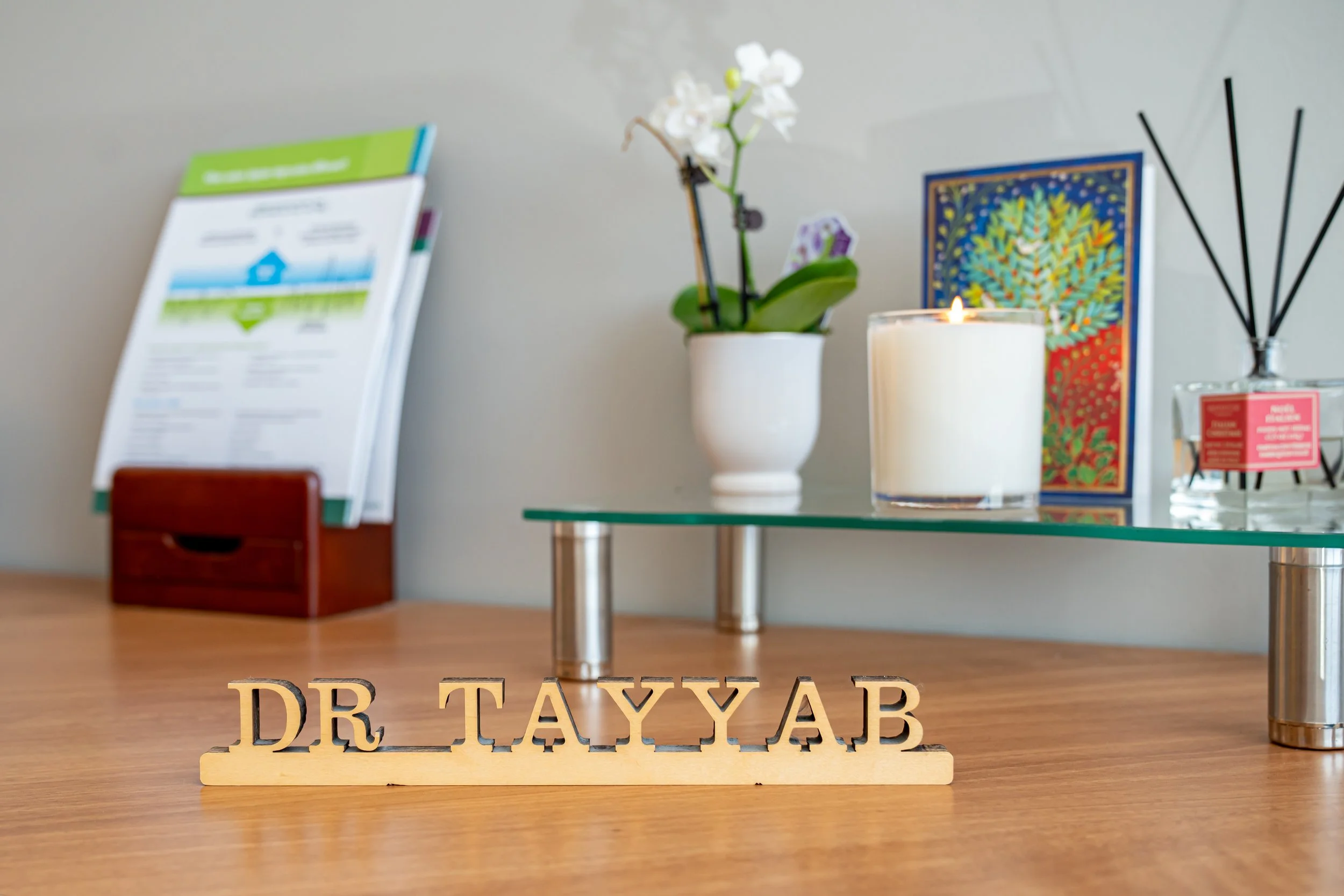 Wooden sign with the name 'Dr. Təyjab' on a wooden desk, behind which is a glass shelf with a white candle, an orchid plant, a colorful artwork, a diffuser, and informational material.