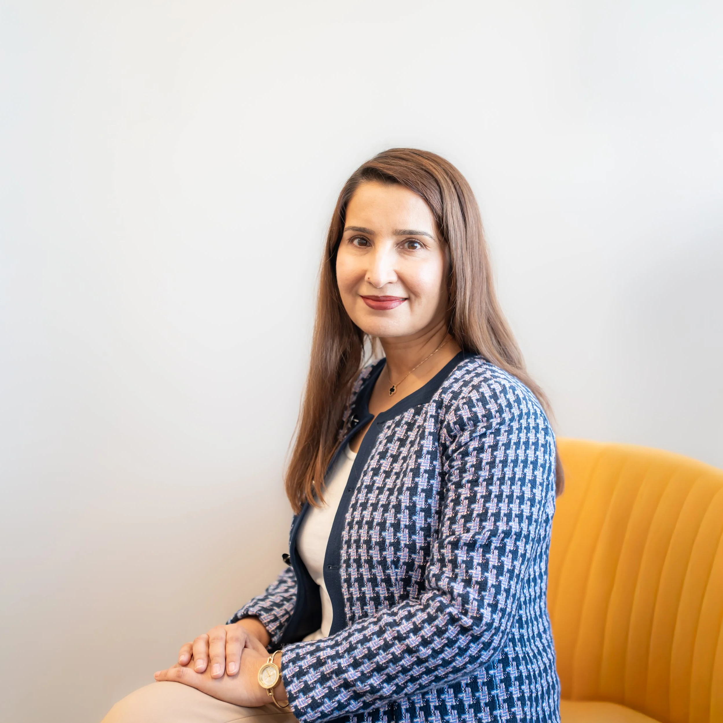A woman with long brown hair, wearing a patterned blazer and white top, sitting on a yellow chair against a plain white wall.