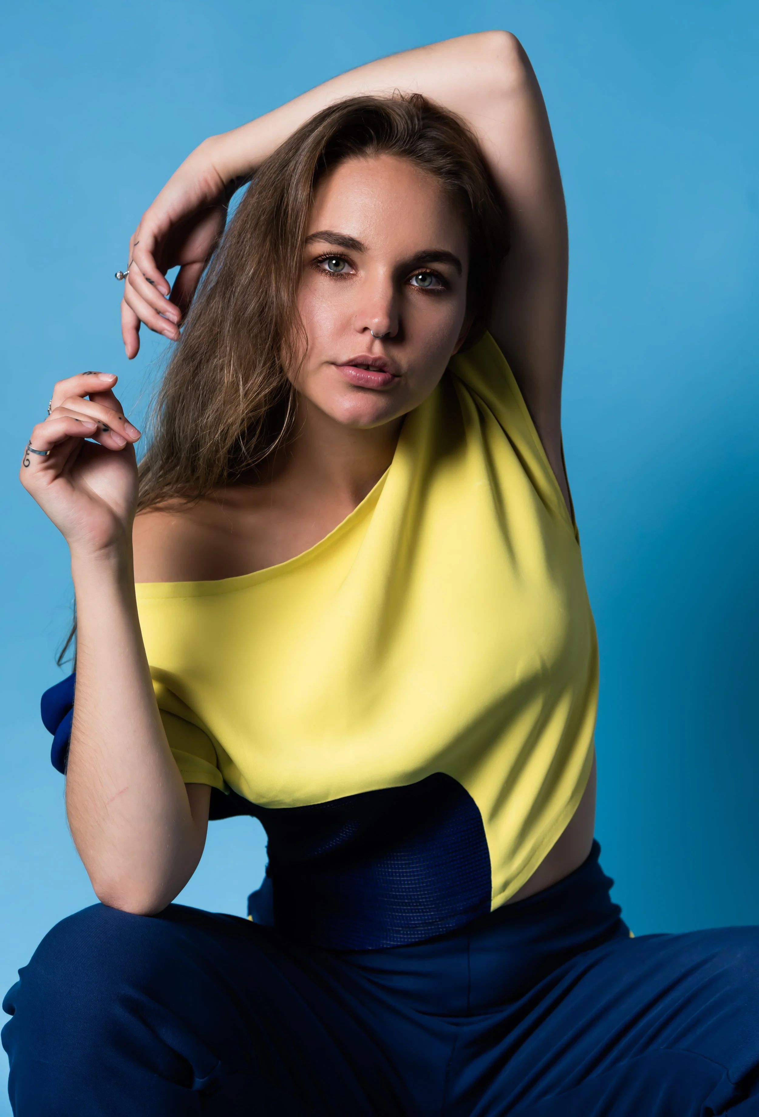 A young woman with long brown hair and blue eyes posing against a blue background, wearing a yellow top and dark pants.