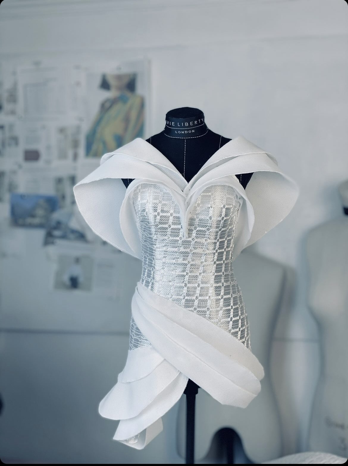 A white sculptural dress on a black dress form with a background of white walls and a blurred artistic setting.